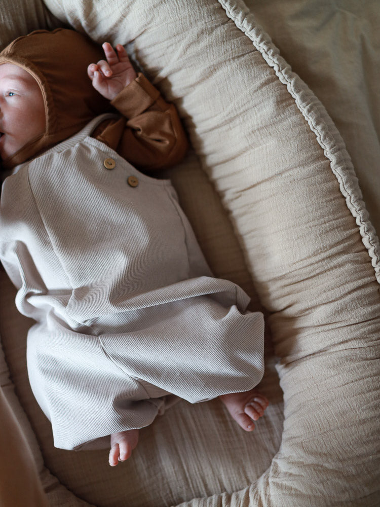 Ein Baby liegt in einem beigen Baby-Nest, trägt eine braune Mütze und die Kurze Baby Schlupflatzhose Feincord - Sand, eine weiche, handgenähte Latzhose aus Feincord ohne Verschlüsse.