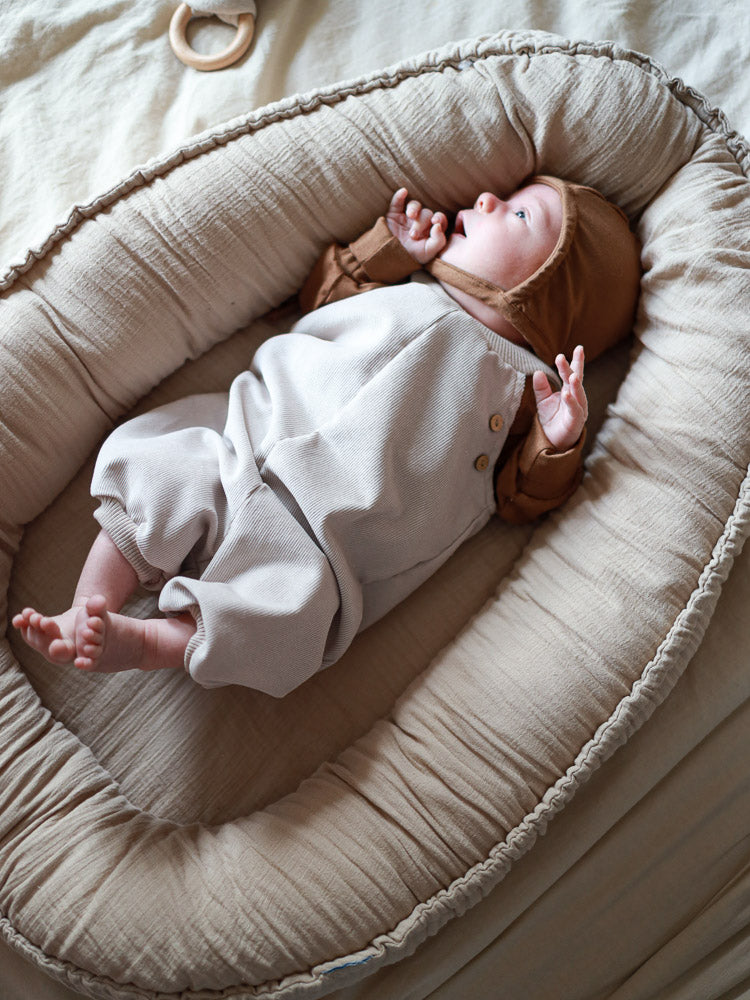 Neugeborenes in einem beigen Nest, trägt eine Kurze Baby Schlupflatzhose Feincord - Sand. Die Latzhose hat Holzknöpfe, weichen Baumwollstoff, ideal zum einfachen Anziehen. Ein Holzring liegt daneben.