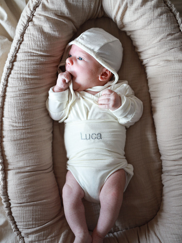 Neugeborenes in einem Individuelle Stickerei off-white Strampler mit Namen Luca, liegend in einem beigen Babynest. Einzigartiges, handgemachtes Geschenk von petit cochon.