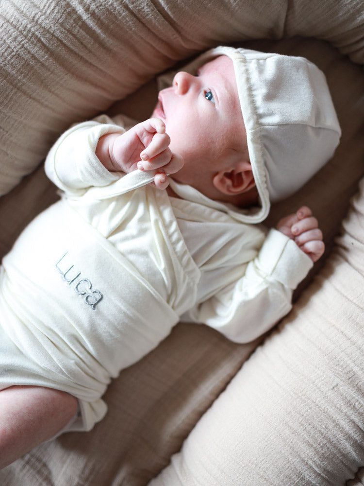 Neugeborenes auf beigem Kissen, trägt einen cremefarbenen Wickelbody mit dem Namen Luca bestickt. Individuelle Stickerei bietet personalisierte, handgemachte Geschenke, perfekt für besondere Anlässe.