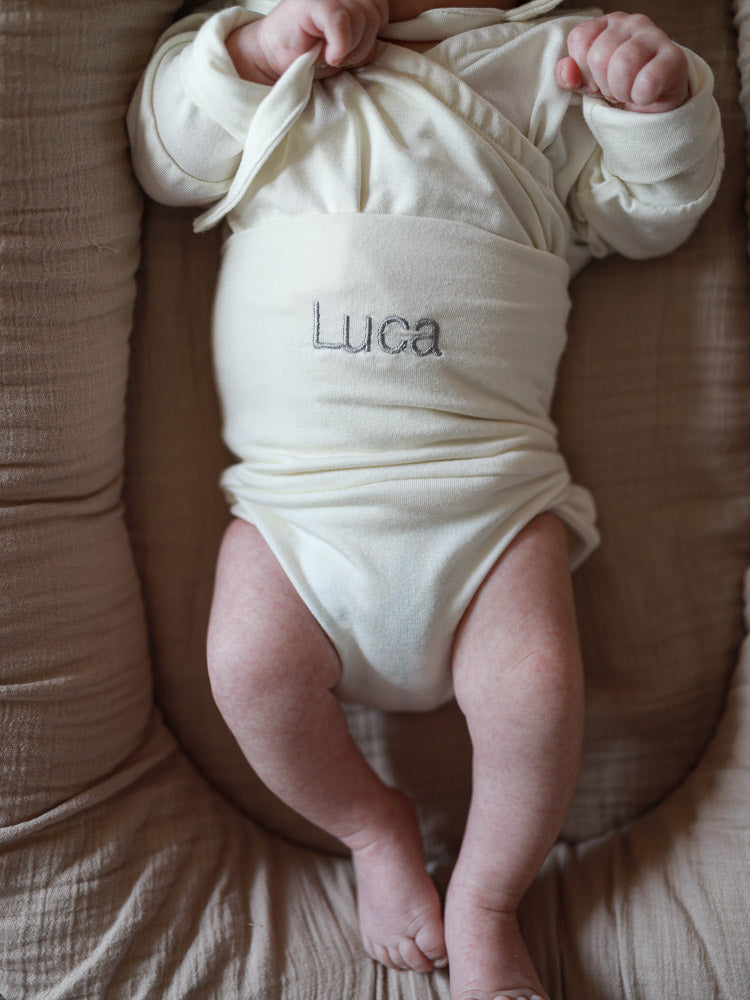 Ein Baby liegt auf einem neutralen Kissen, trägt einen cremefarbenen Einteiler mit Luca aufgestickt. Individuelle Stickerei macht es zu einem einzigartigen, personalisierten Geschenk.