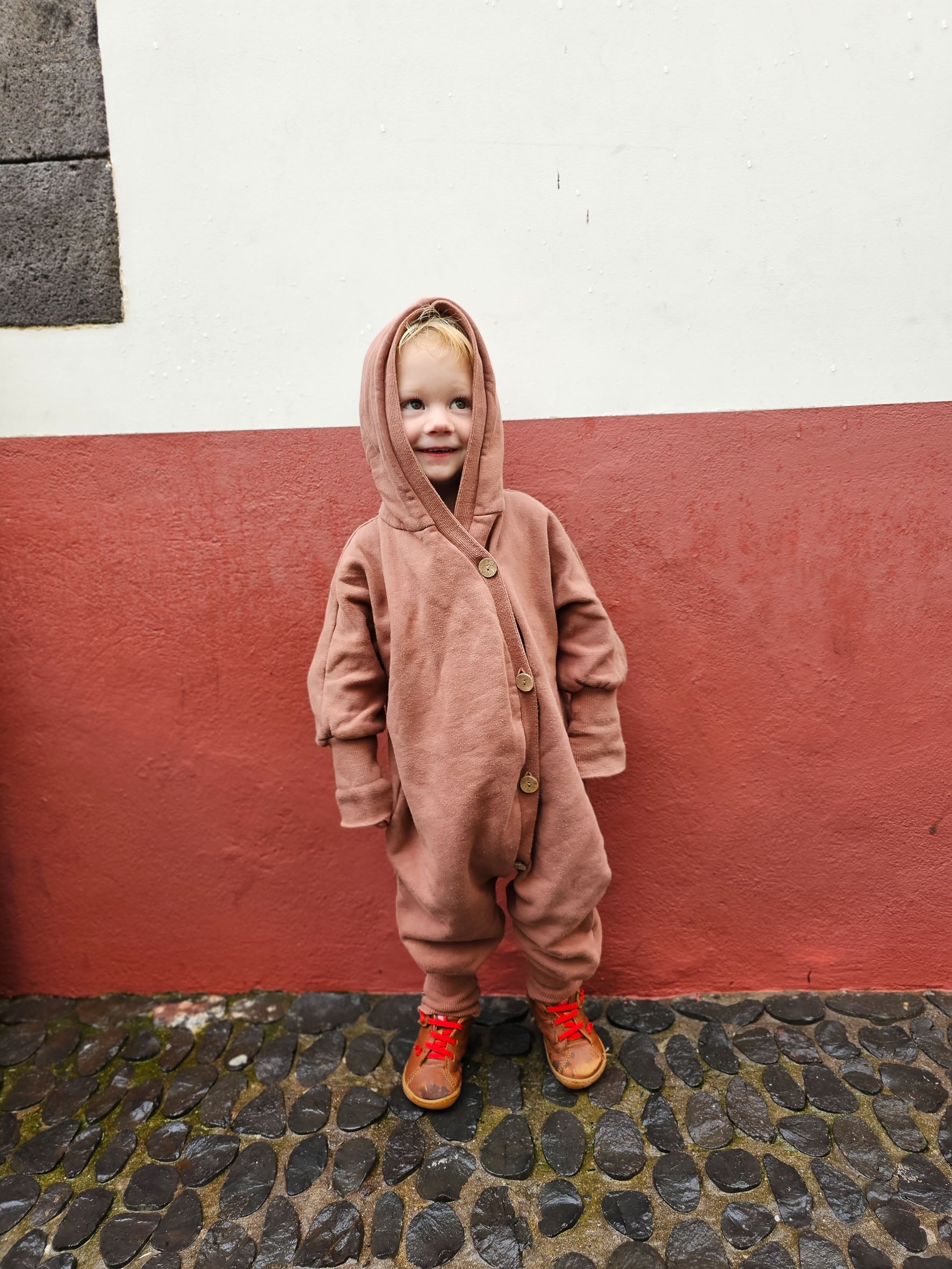 Kind trägt den Kuschelanzug Bio-Sweat - Zimt mit Wichtelkapuze, aus weichem Bio-Baumwolle-Sweat, ideal für drinnen und draußen. Perfekt für Komfort und Wärme, mit mitwachsendem Schnitt.