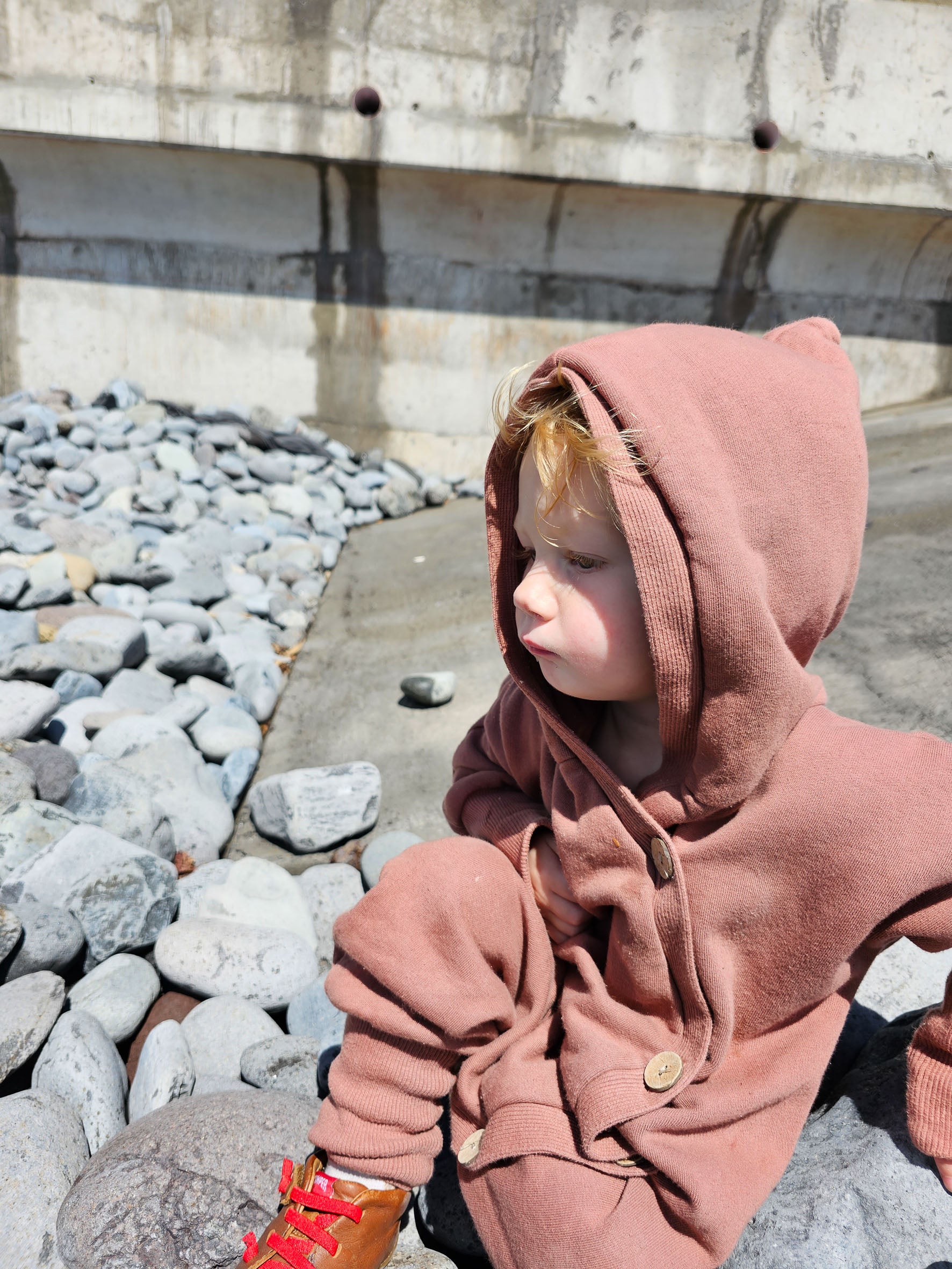 Kleinkind sitzt auf Felsen, trägt Kuschelanzug Bio-Sweat - Zimt mit Wichtelkapuze. Der Overall wirkt gemütlich und locker, ideal für Outdoor-Aktivitäten oder als wärmende Schicht an kühleren Tagen.