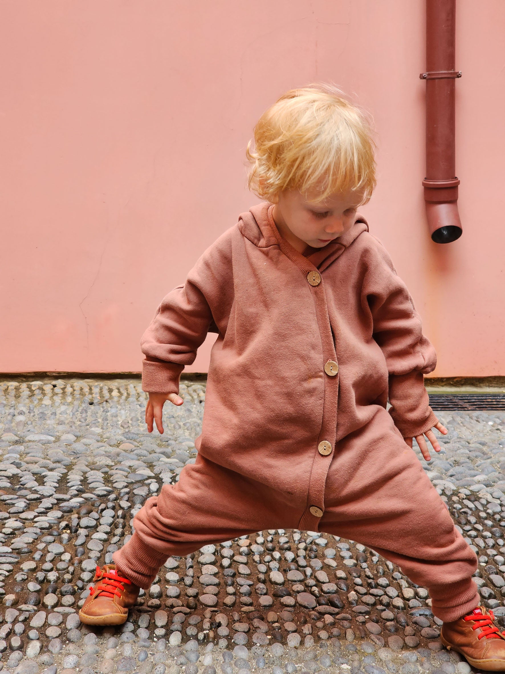 Kind trägt Kuschelanzug Bio-Sweat - Zimt mit Wichtelkapuze; Overall aus Bio-Baumwolle, ideal für drinnen und draußen.