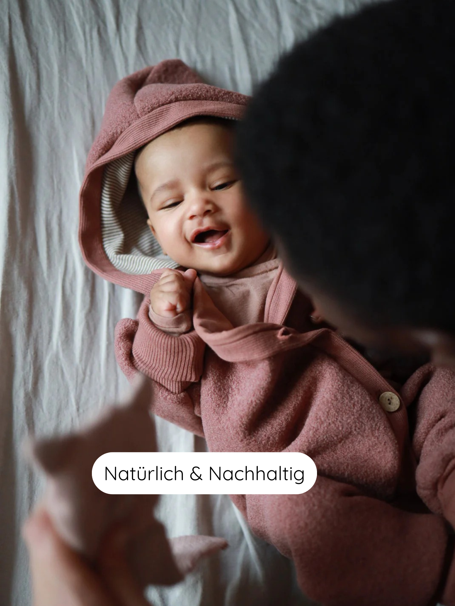 Ein lächelndes Baby liegt auf einem hellen Laken, gekleidet in einen Kuschelanzug Wollwalk - Zimt mit Kapuze, während eine Hand ein kleines Stofftier hält. Das Label „Natürlich & Nachhaltig“ ist sichtbar.