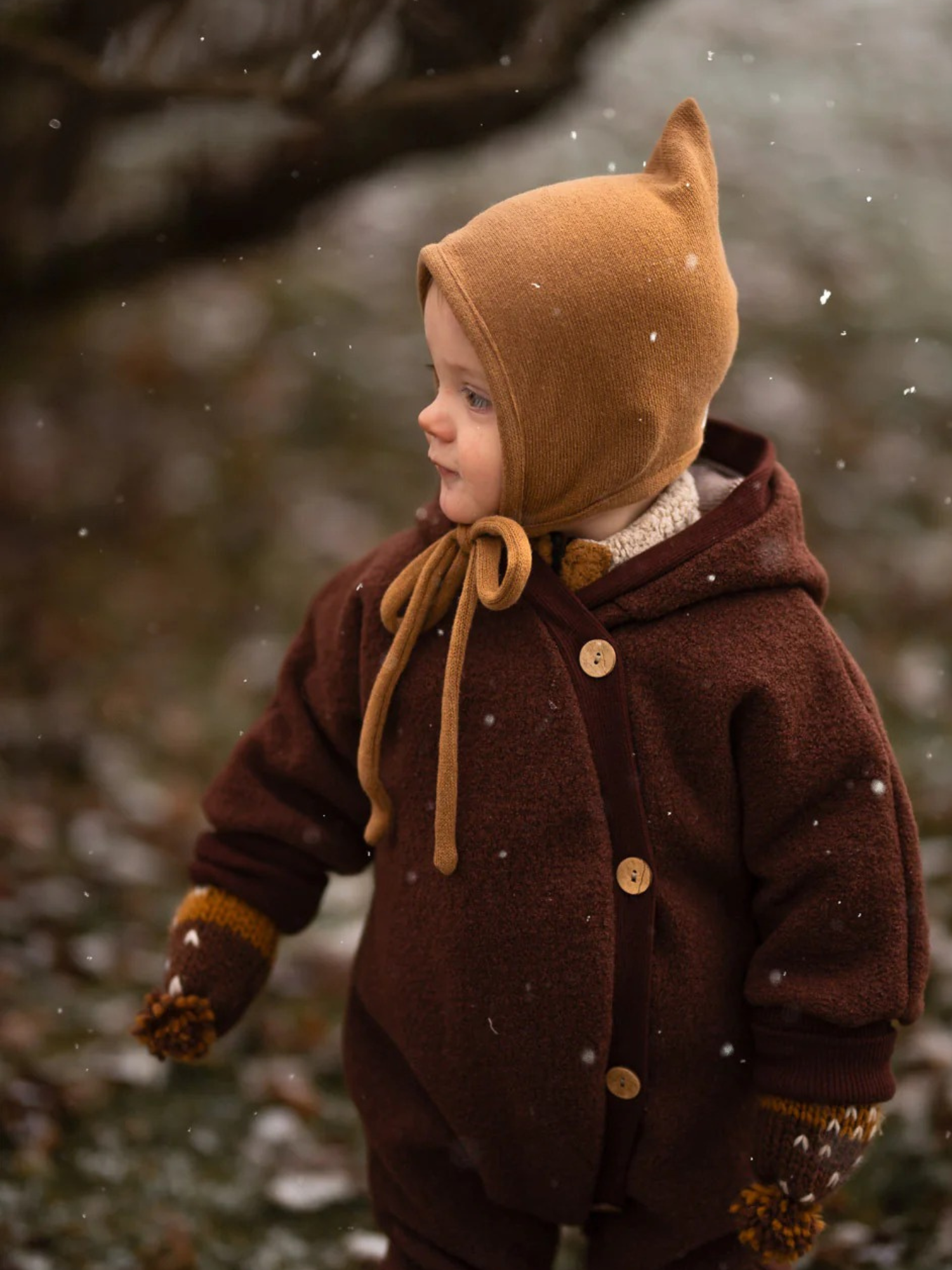 Ein Kind trägt den Kuschelanzug Wollwalk - Schoko, einen dunkelbraunen Einteiler aus 100% Schurwolle mit Bio-Jersey-Futter, passend für kalte Tage. Ergänzt mit passenden Strickfäustlingen und gelber Mütze.
