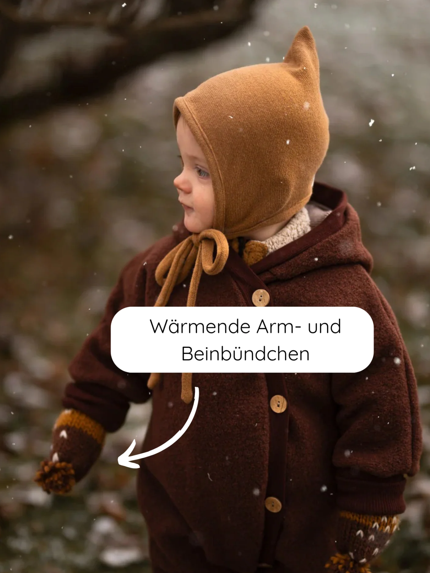 Ein kleines Kind im Freien trägt den Kuschelanzug Wollwalk - Schoko, komplett mit einem passenden, gestrickten Bonnet und Fäustlingen, während ein Pfeil auf die wärmenden Arm- und Beinbündchen zeigt.