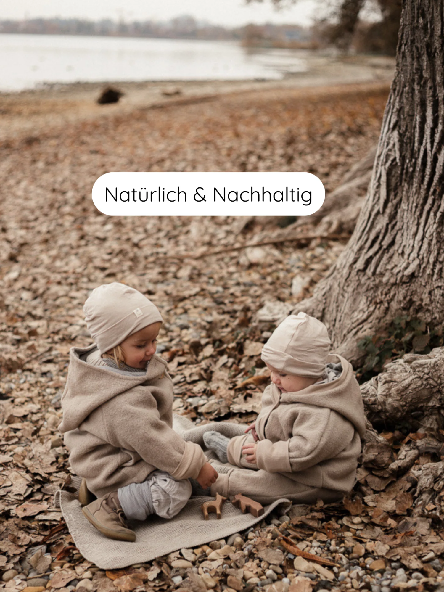 Zwei Kleinkinder in Kuschelanzug Wollwalk - Sand spielen mit Holzfiguren auf einer Decke am See. Die Anzüge aus 100% Schurwolle bieten Bewegungsfreiheit und sind mit Baumwollfutter ausgestattet.