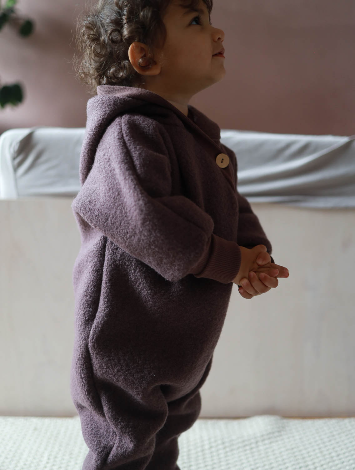 Kleinkind im Raum stehend, trägt den Kuschelanzug Wollwalk - Malve aus 100% Schurwolle mit Bio-Jersey-Futter, ideal für Jahreszeiten und hält warm, mit langen Bündchen für optimalen Schutz. Wollwalk Anzug, Wollwalk Overall für Kinder,