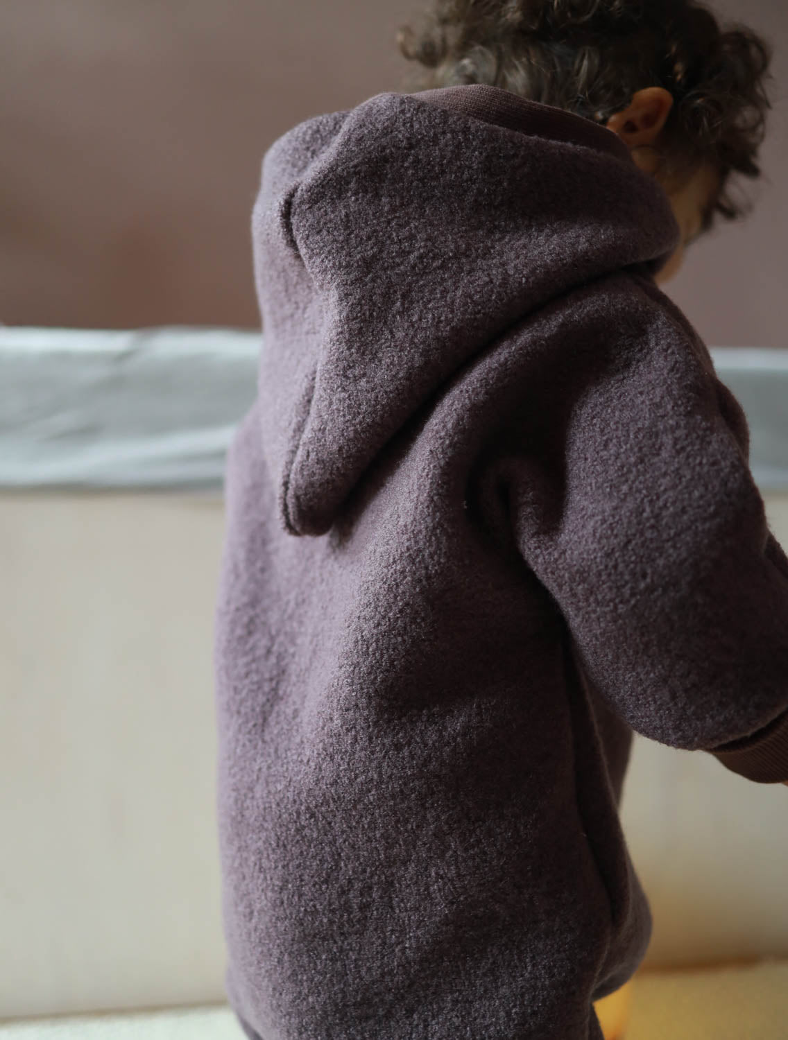 Kleinkind trägt den Kuschelanzug Wollwalk - Malve mit eng anliegenden Bündchen, ideal für kalte Tage. Der Overall hat eine Wichtelkapuze und bietet Bewegungsfreiheit, perfekt für den Herbst und Winter. Wollwalk Anzug, Wollwalk Overall für Kinder