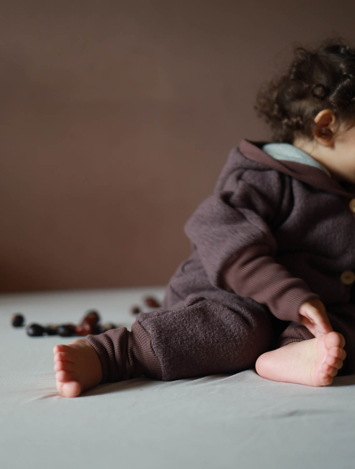 Ein Baby sitzt auf einem Bett, bekleidet mit dem Kuschelanzug Wollwalk - Malve. Der Overall hat enganliegende Arm- und Beinbündchen und bietet Bewegungsfreiheit durch den tiefen Schritt. Wollwalk Anzug, Wollwalk Overall für Kinder, mitwachsender Wollwalk