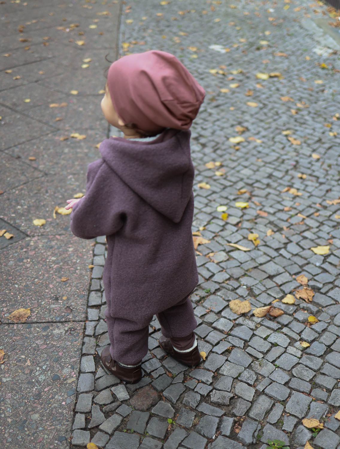 Kind trägt den Kuschelanzug Wollwalk - Malve aus Schurwolle, steht auf einem Ziegelweg. Der Overall hat eine Wichtelkapuze, enge Bündchen und bietet Bewegungsfreiheit und Schutz vor Wind. Wollwalk Anzug, Wollwalk Overall für Kinder, mitwachsender Wollwalk