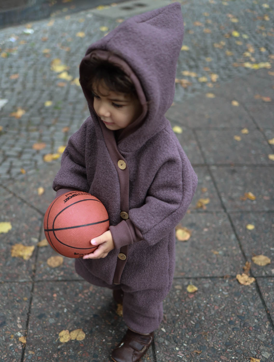 Ein Kind hält einen Basketball und trägt den Kuschelanzug Wollwalk - Malve, ideal für kalte Tage, mit enganliegenden Bündchen und Kapuze, um warm und geschützt zu bleiben. Wollwalk Overall für Kinder und Babys, made in Berlin