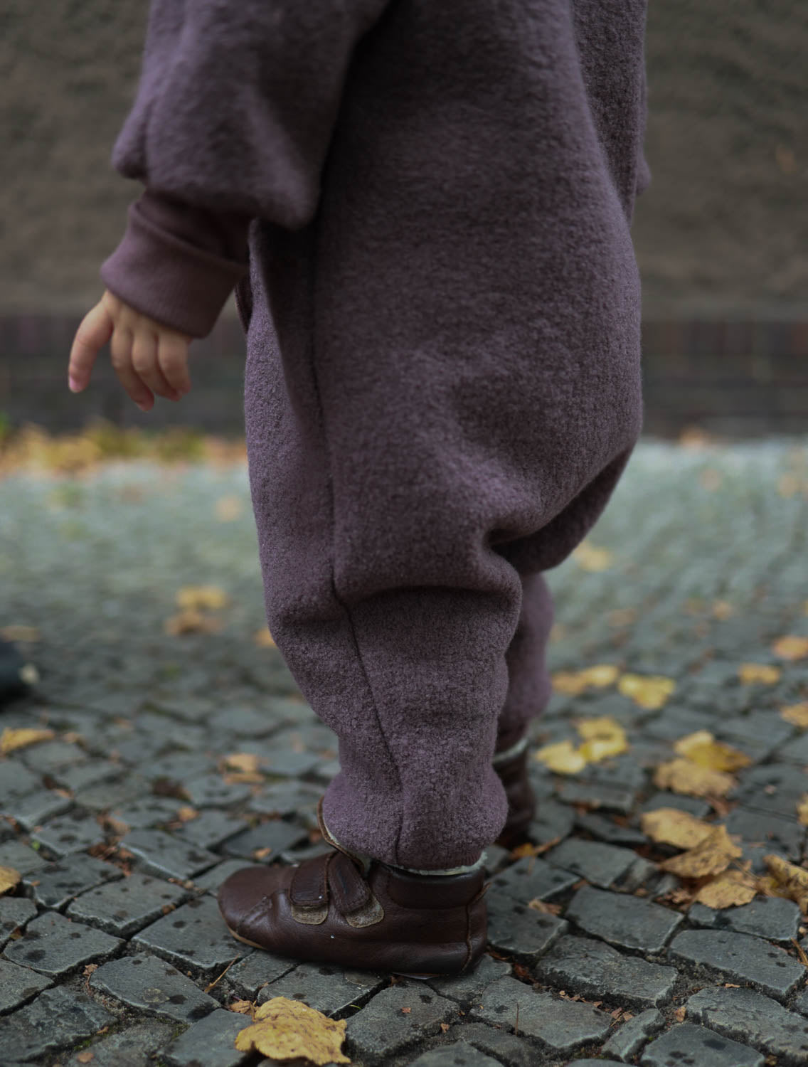 Kleinkind trägt Kuschelanzug Wollwalk - Malve. Sichtbar sind Beine mit lila Outfit und braunen Schuhen auf Ziegelboden. Ideal für kühle Jahreszeiten, hält warm und bietet Bewegungsfreiheit. Wollwalk Anzug, Wollwalk Overall für Kinder