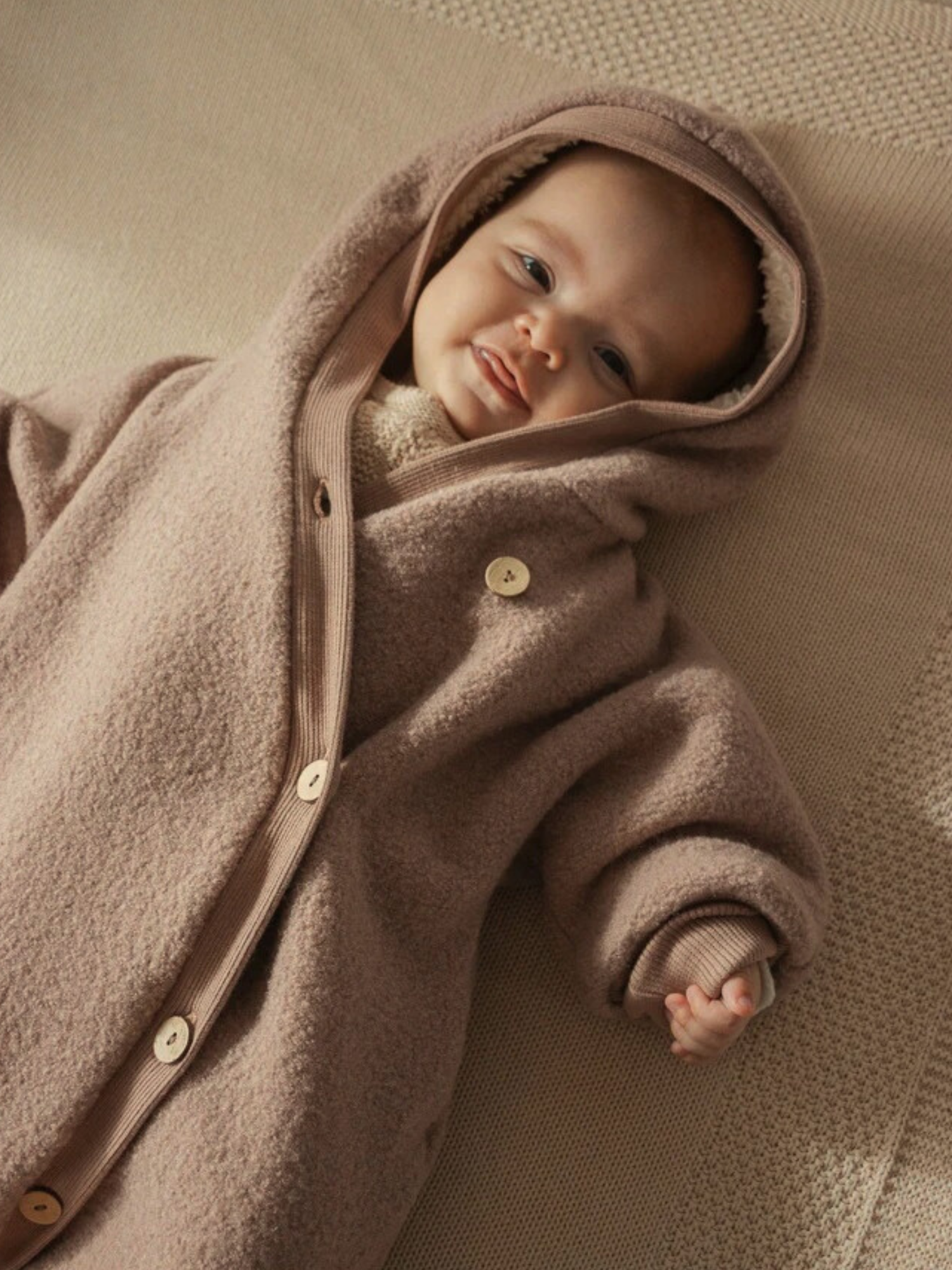 Winter Wollwalk Overall mit Teddyfutter- Puder: Ein Baby in einem beigen, kuscheligen Wollwalk-Overall mit Kapuze und großen Knöpfen, liegend auf einer weichen Oberfläche, zeigt ein zufriedenes Gesicht und eine kleine Hand.