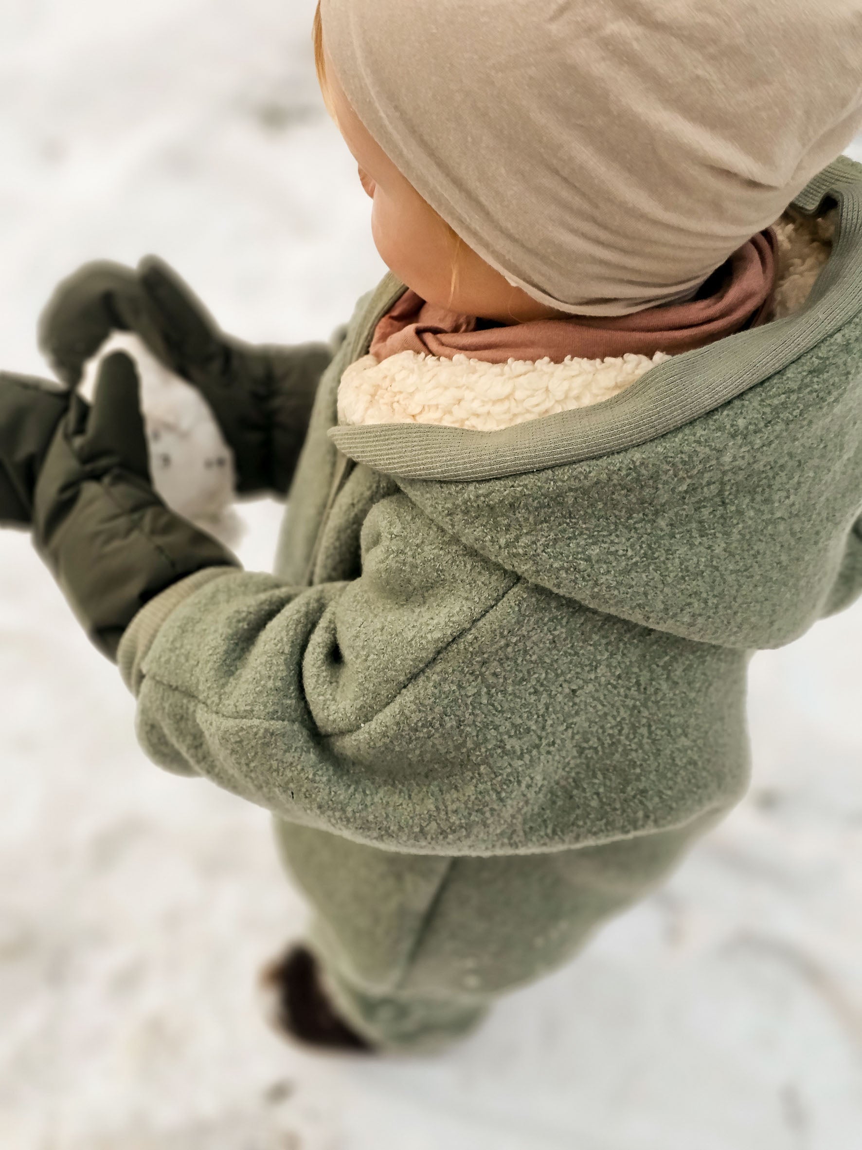 Kind spielt im Schnee, trägt den Kuschelanzug Wollwalk - Pistazie mit passender Mütze. Der Schneeanzug für Kinder bietet durch seine Bündchen Schutz vor Kälte und ist ideal für kalte Wintertage. Wollwalk Anzug für Kinder und Babys