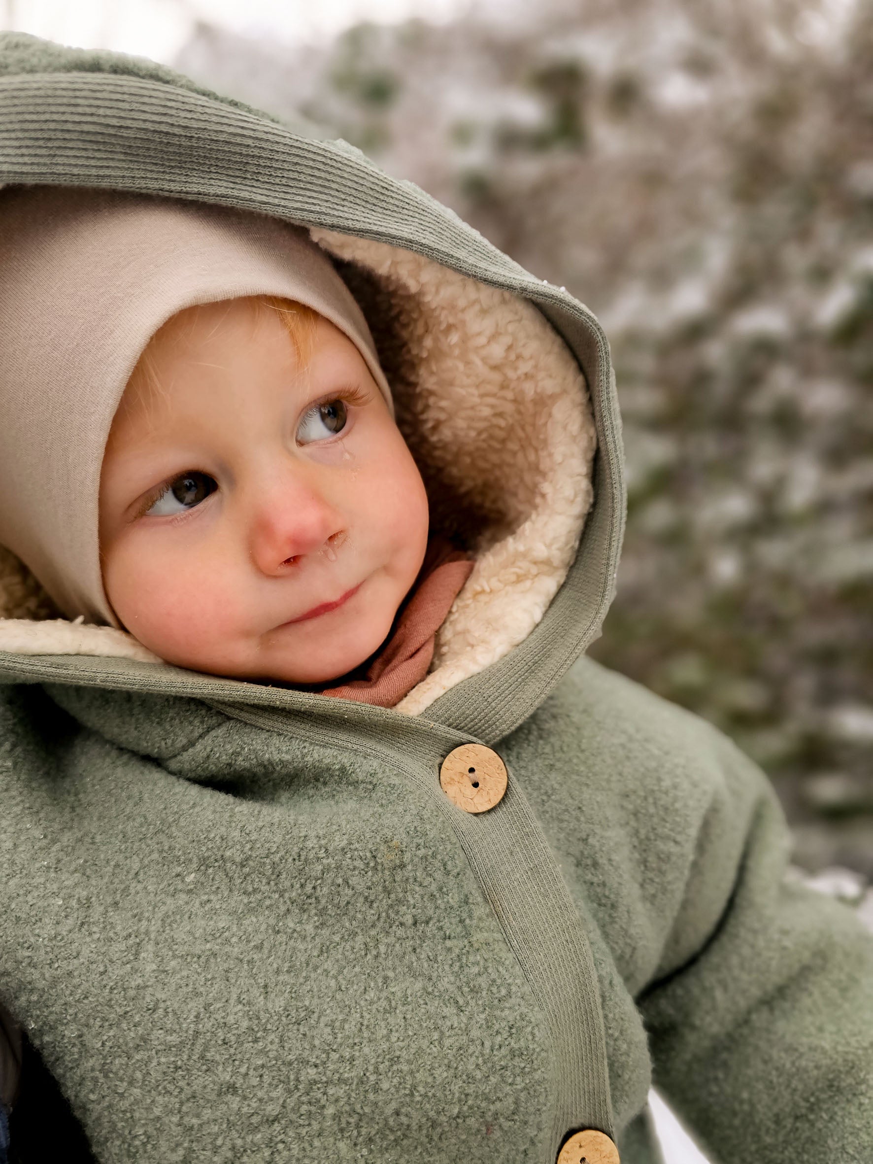 Kuschelanzug Wollwalk - Pistazie: Ein Baby trägt einen grünen Wollwalk Overall mit Kapuze, ideal für kühle Jahreszeiten. Der Schneeanzug bietet Wärme und Bewegungsfreiheit, perfekt für draußen und Babytragen. Wollwalk Anzug für Kinder und Babys