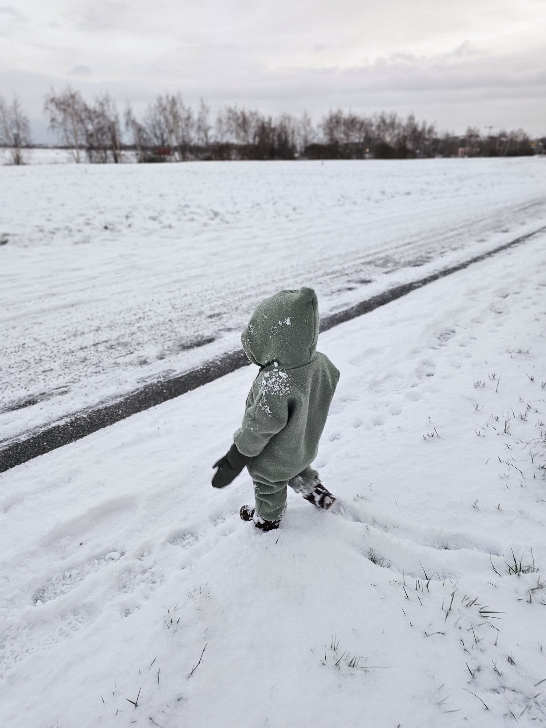 Kind im Schnee, trägt den Kuschelanzug Wollwalk - Pistazie aus 100% Schurwolle, ideal für kalte Jahreszeiten mit wärmenden und atmungsaktiven Eigenschaften.
