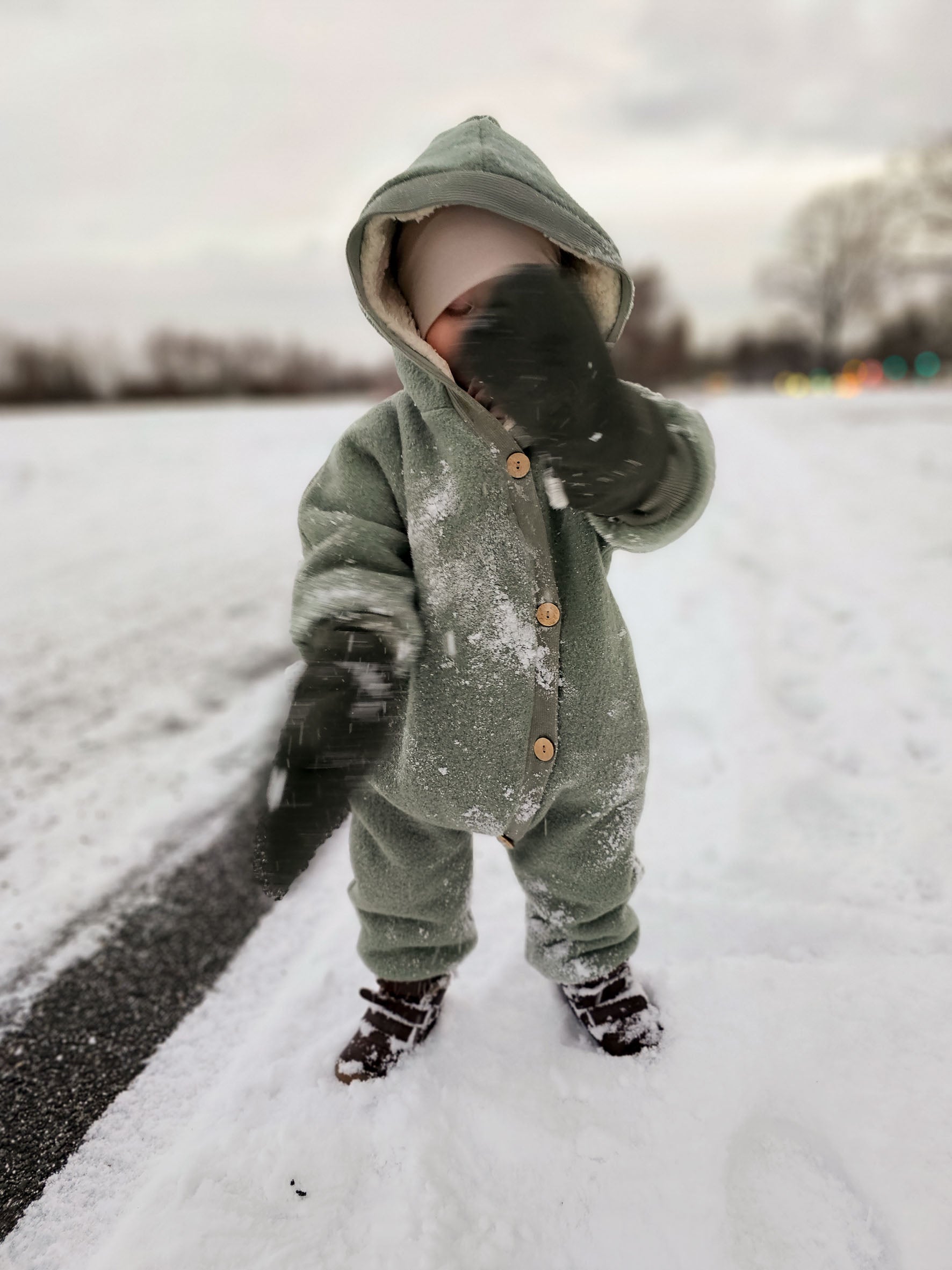 Kind im Kuschelanzug Wollwalk - Wollwalk Overall in Grün, spielt im Schnee; Overall aus Schurwolle mit Bio-Jersey-Futter, bietet Wärme und Schutz, ideal für kalte Jahreszeiten.