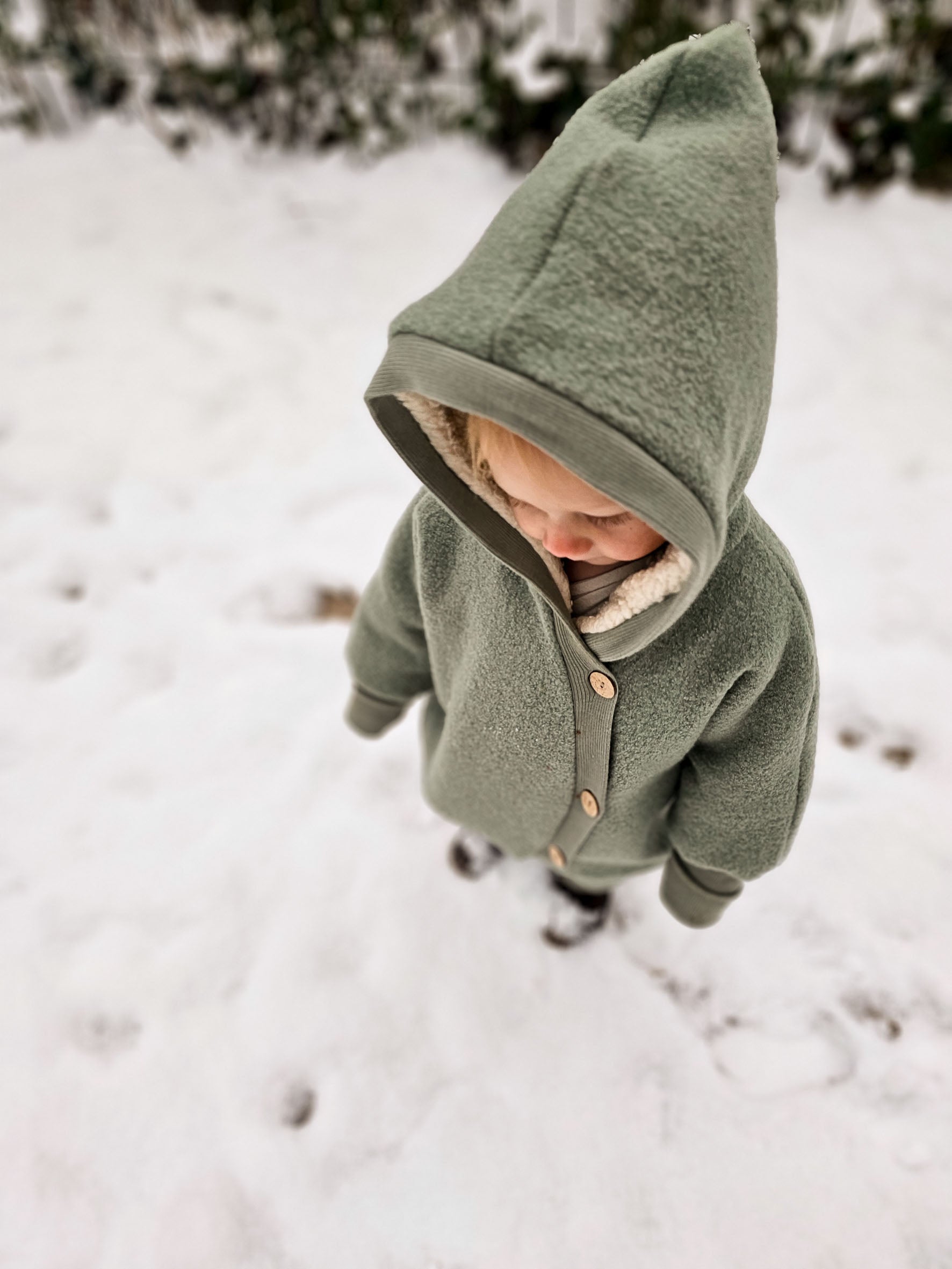 Ein Kind steht im Schnee und trägt den Kuschelanzug Wollwalk - Pistazie mit einer Kapuze, ideal für kalte Tage. Der Anzug schützt vor Kälte mit eng anliegenden Bündchen. Wollwalk Anzug für Kinder und Babys