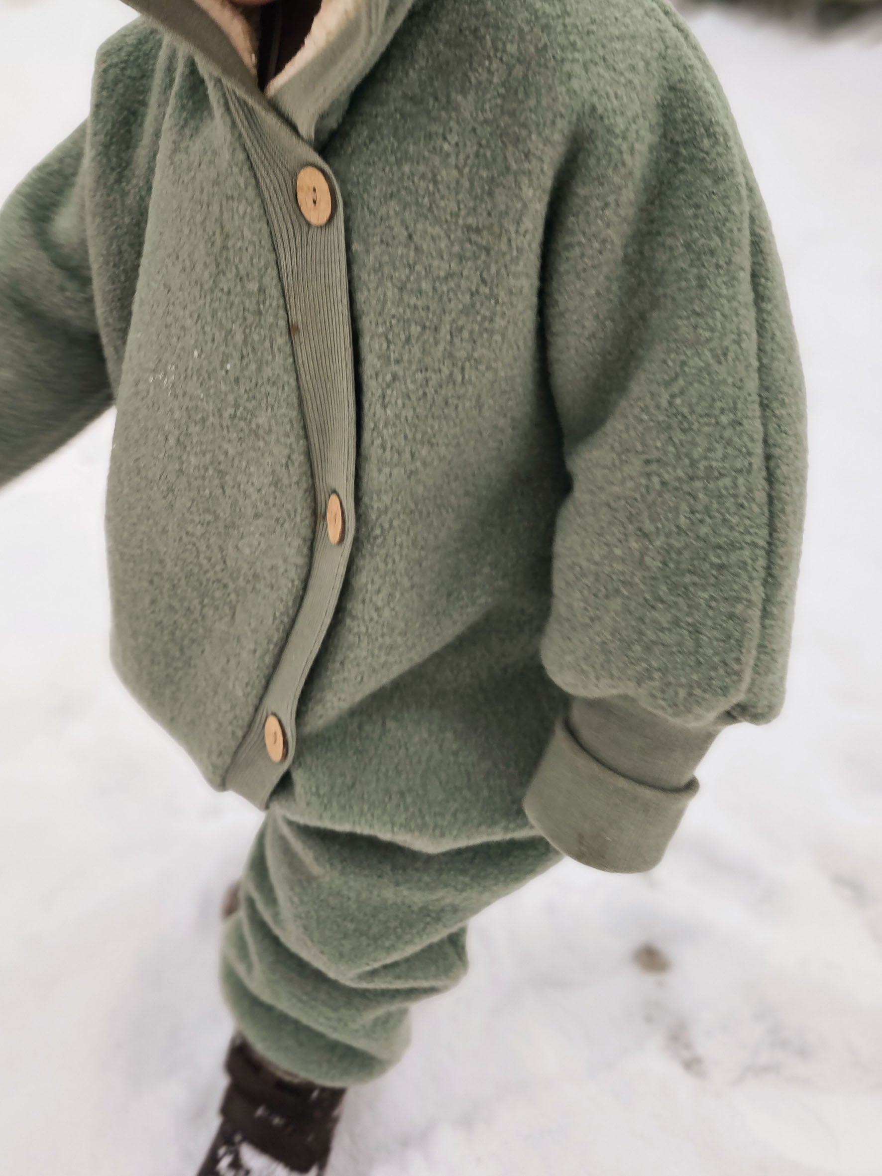 Kuschelanzug Wollwalk - Pistazie: Ein Kind trägt einen grünen Wollwalk Overall mit langen Bündchen und Kapuze, ideal für kalte Temperaturen und Babytragen, bietet Wärme und Bewegungsfreiheit. Wollwalk Anzug für Kinder