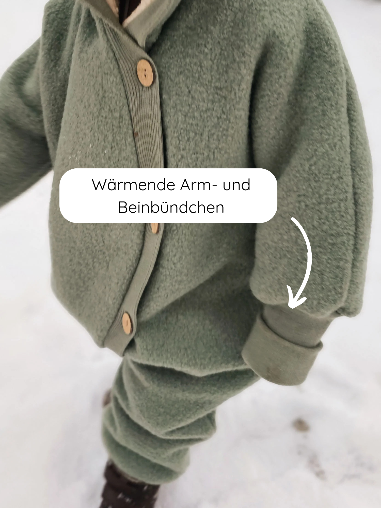 Kleinkind im Winter Wollwalk Overall mit Teddyfutter - Pistazie, trägt eine olivgrüne Fleecejacke mit Holzknöpfen und passenden Hosen, steht im Schnee. Ribbelbündchen an Ärmeln und Beinen bieten Schutz.