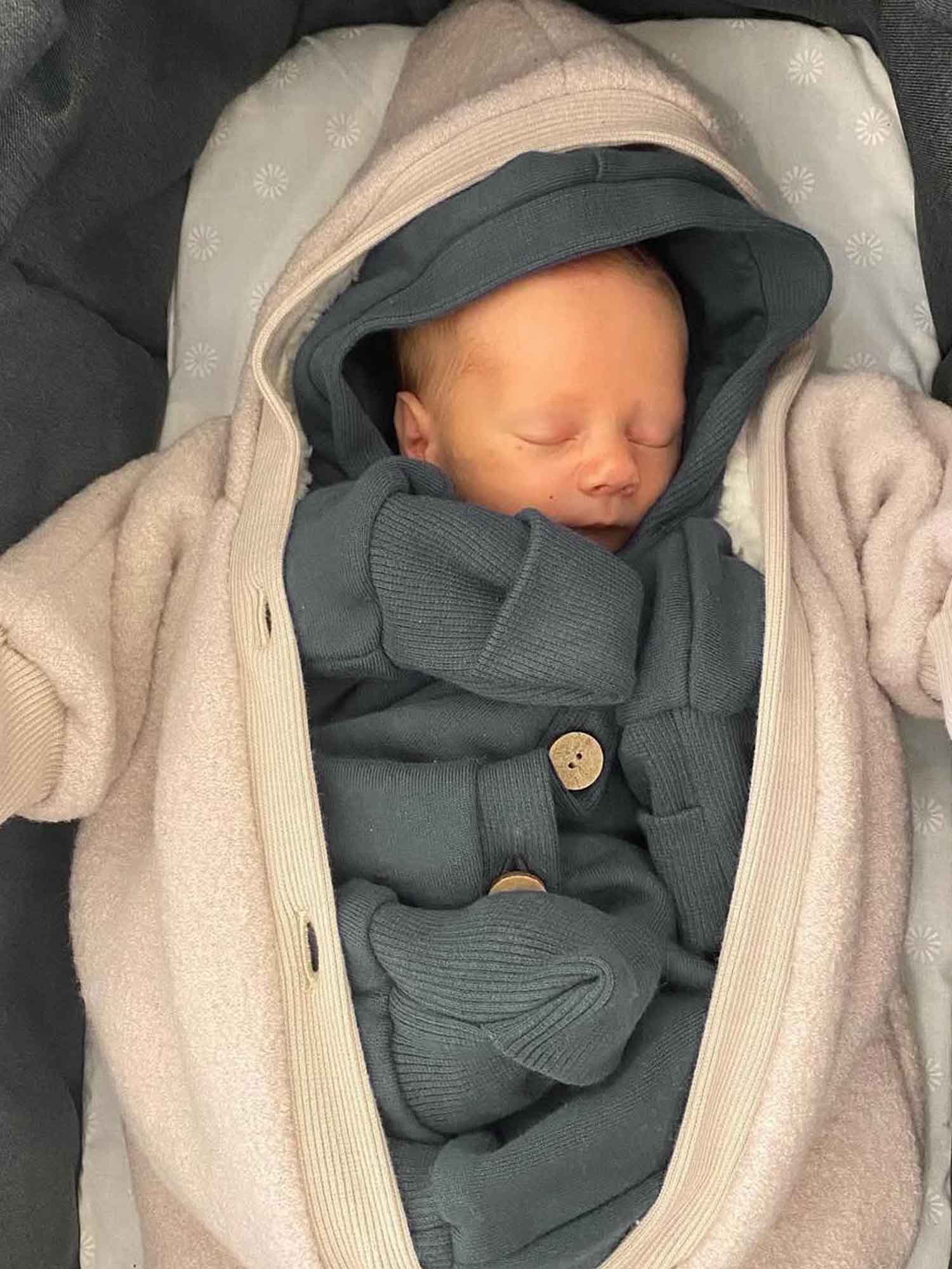 Neugeborenes schläft im Teddy-Kuschelanzug Wollwalk - Sand, kuschelig verpackt in einem weichen, gefütterten Overall mit Kapuze und Fäustlingen, in einem Kinderwagen-Einsatz gebettet.