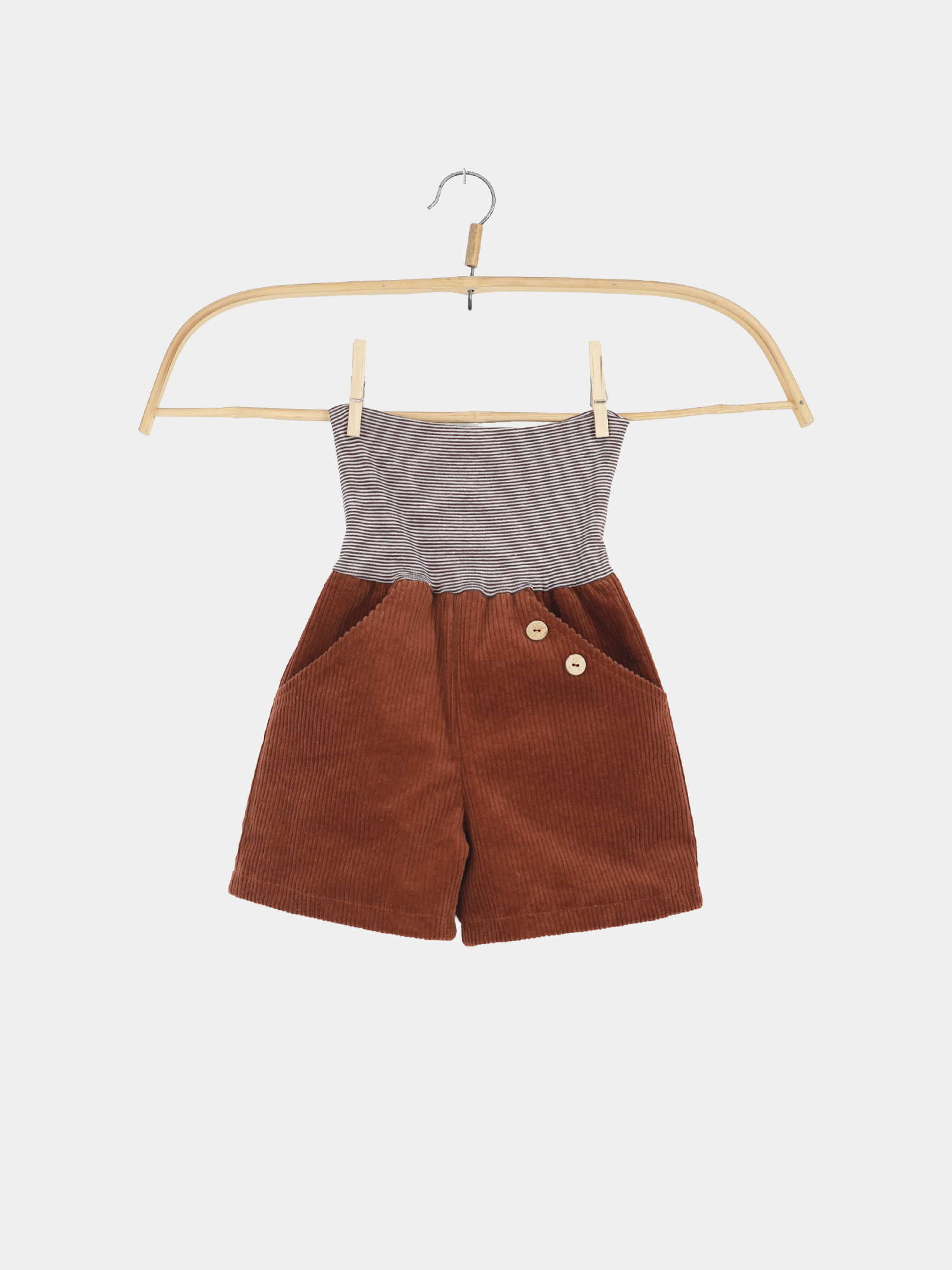 Kurze Buxe Breitcord - Rost: Ein Kinderstrampler mit grauem Ringel-Oberteil und rostbraunem Kordunterteil, zwei Fronttaschen und seitlichen Holzknöpfen, präsentiert auf einem Holzbügel.