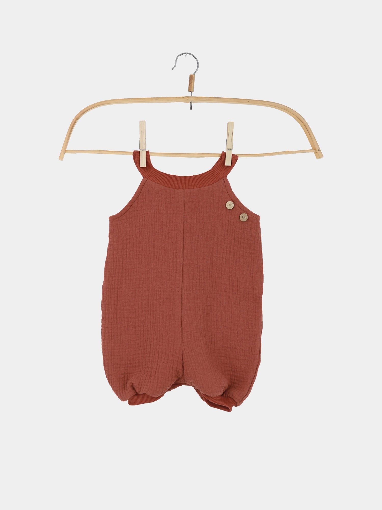 Kurze Schlupflatzhose Bio-Musselin - Terrakotta: Handgenähter Kinderromper aus strukturiertem Stoff mit zwei Holzknöpfen an der Schulter, präsentiert auf einem Holzkleiderbügel, ideal für bequeme Sommerabenteuer.