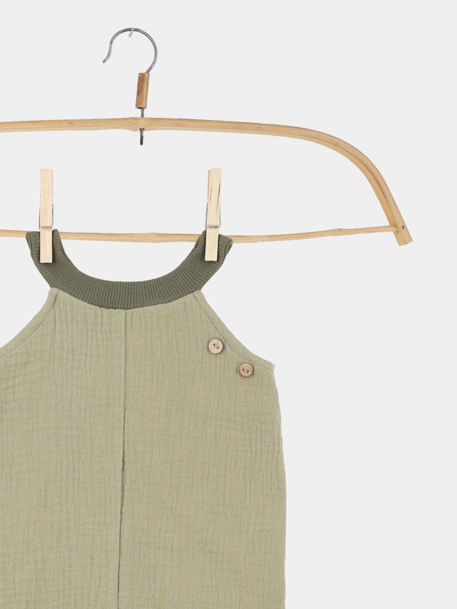 Kurze Schlupflatzhose Musselin - Salbei: Ein ärmelloser, strukturierter Romper für Kleinkinder, aus Musselin. Er hängt an einem Holzkleiderbügel, mit zwei kleinen Holzknöpfen auf einer Schulter.