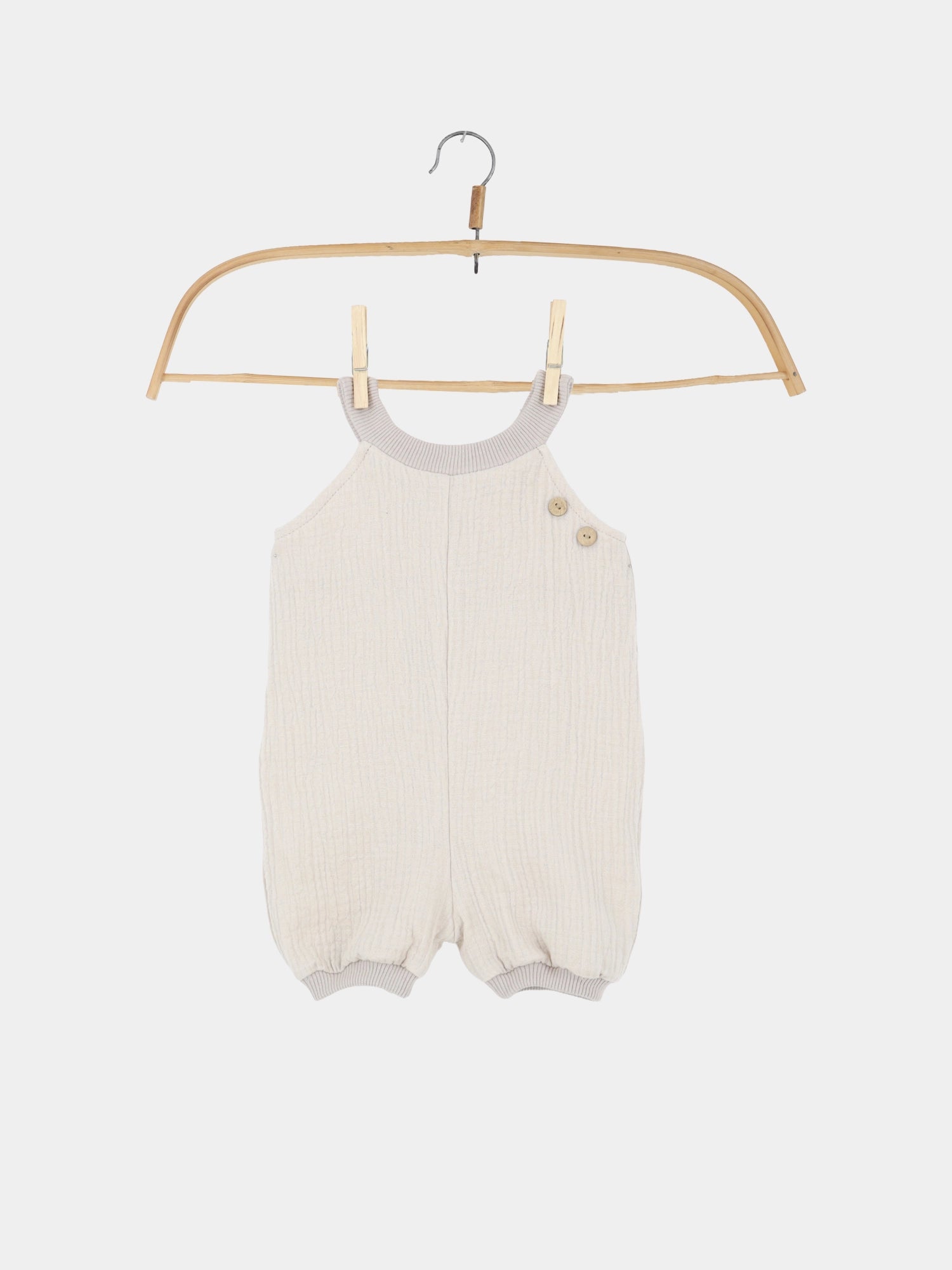 Kurze Schlupflatzhose Bio-Musselin - Sand: Ein cremefarbener, ärmelloser Baby-Strampler aus weichem Musselin mit geripptem Ausschnitt, Beinbündchen und zwei kleinen Holzknöpfen auf einer Schulter. Ideal für den Sommer.