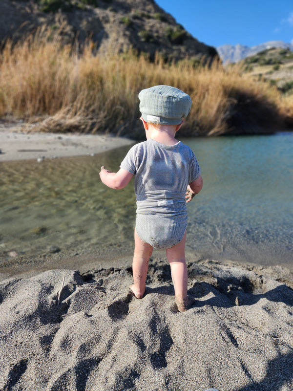 Ein Kleinkind steht am Ufer in einem Loopbody Kurzarm Bio-Baumwolle - Ringel Maus, der einen weichen, gestreiften Stoff zeigt, ideal für sommerliche Temperaturen.