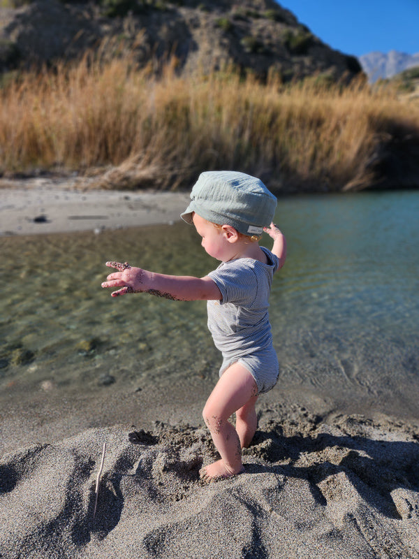 Ein Kleinkind spielt am Wasser in einem Kurzarm Loopbody aus Bio-Baumwolle, genannt Ringel Maus, mit Streifenmuster. Sand klebt an den Beinen und Händen des Kindes.