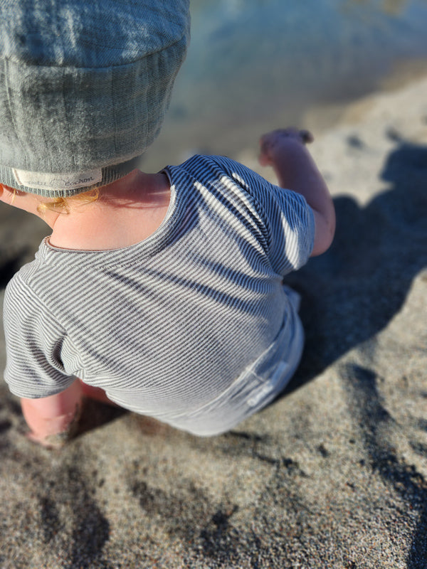 Ein Kleinkind am Wasser trägt einen Loopbody Kurzarm Bio-Baumwolle - Ringel Maus, mit schmalen Streifen und Denim-Bucket-Hut, greift neugierig Richtung Wasser.