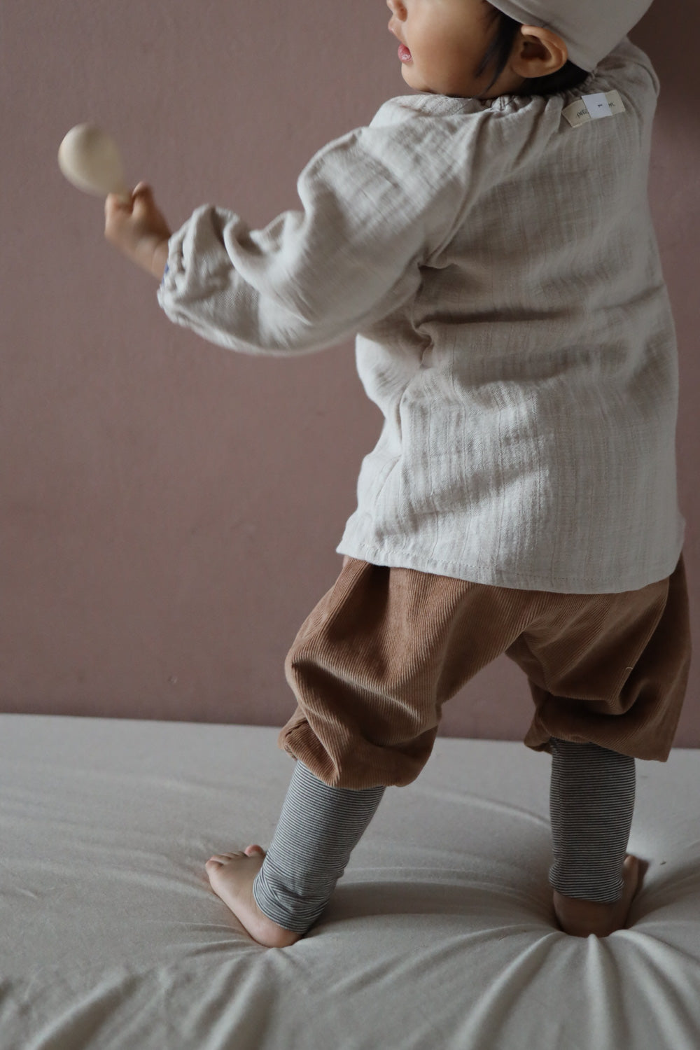 Kleinkind in Knickerbocker Feincord Ringel - Weizen, locker sitzende Cordhose mit gestreiftem Bund, ideal für Bewegungsfreiheit. Das Kind hält ein Holzspielzeug, trägt eine Mütze und Rollkragenpulli.