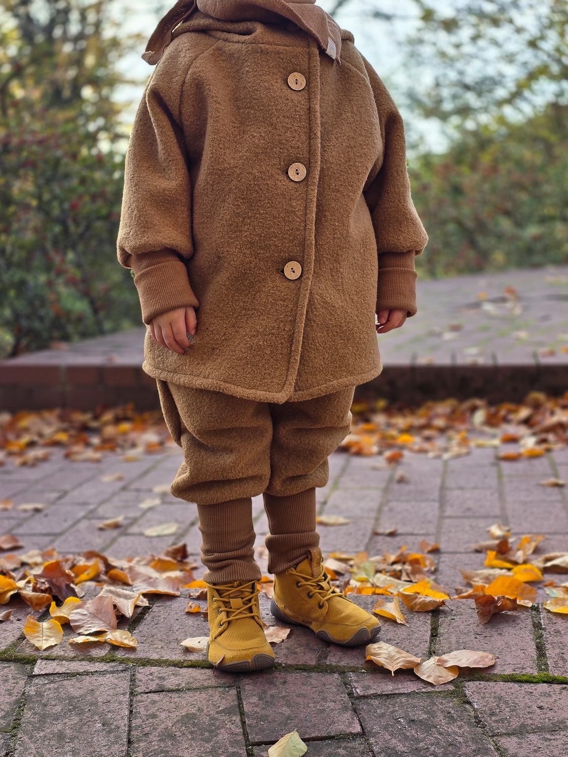 Person trägt Knickerbocker Wollwalk - Kamel, passende Wollhose für kaltes Wetter. Sichtbar sind die Kängurutaschen, das Bio-Baumwollbündchen und die wärmenden Eigenschaften, ideal für Herbstaktivitäten.