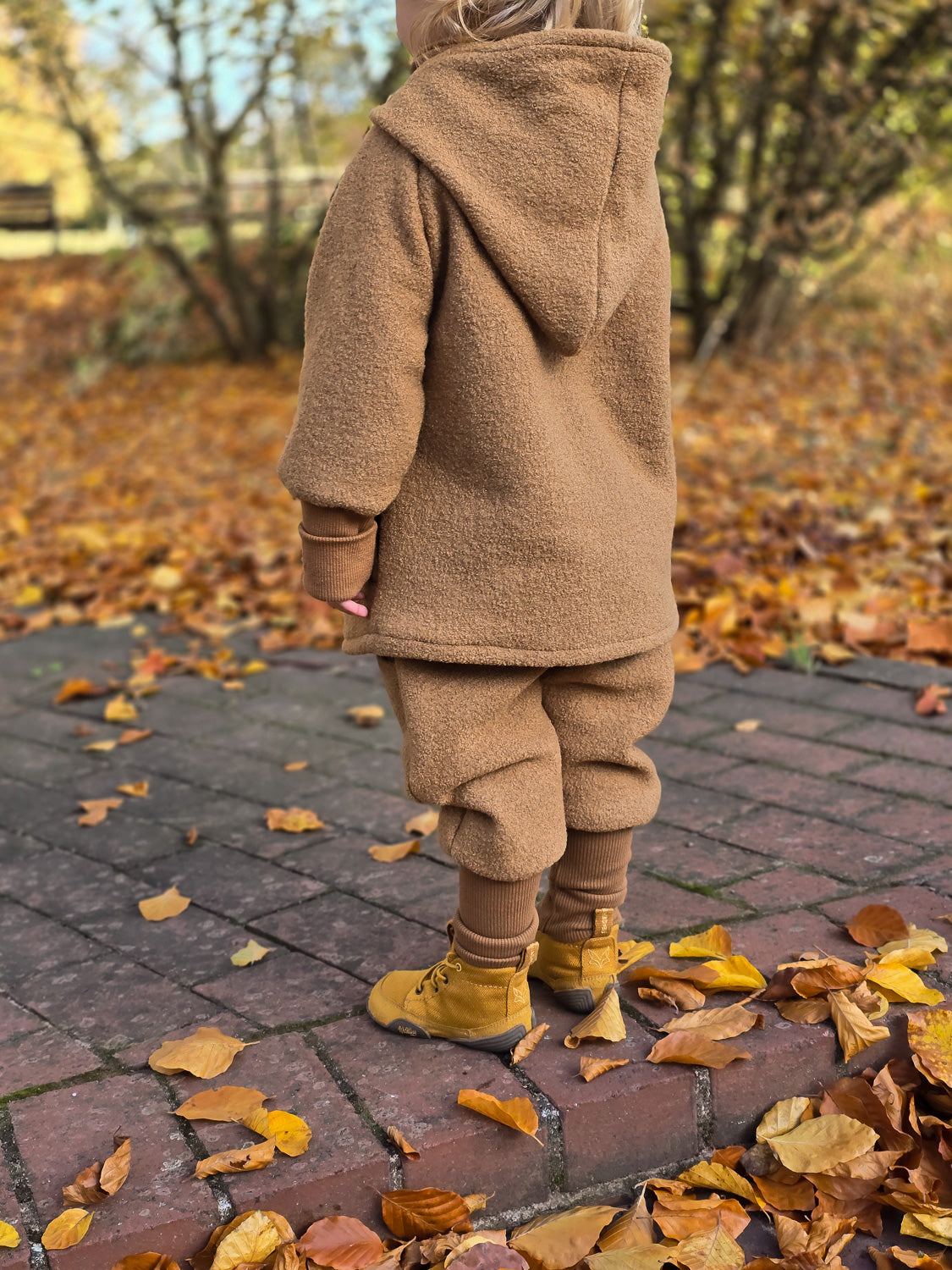 Ein Kind trägt die Knickerbocker Wollwalk - Kamel auf einem Ziegelweg, ideal für kaltes Wetter, mit praktischen Kängurutaschen und krempelbaren Bündchen, perfekt zum Tragen über Leggings oder Strumpfhosen.