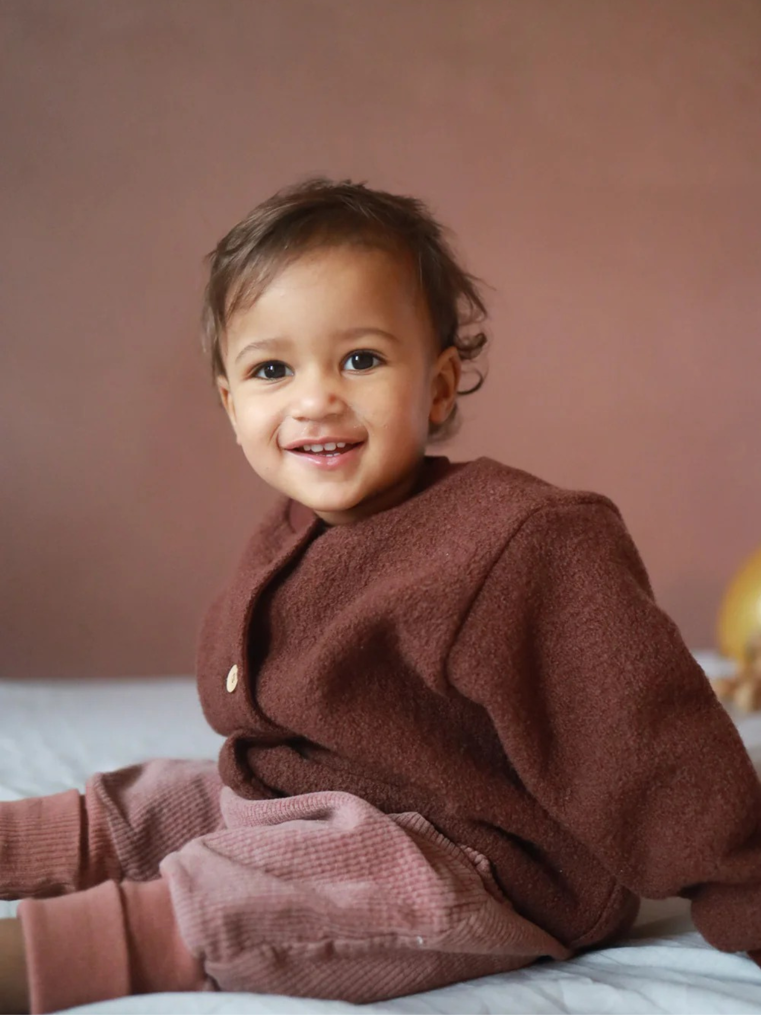 Ein lächelndes Kleinkind trägt die Knickerbocker Breitcord Stretch - Rosenholz, eine weiche, braune Cordhose mit bequemem Schnitt, ideal für kühle Tage und aktive Entdeckungstouren.