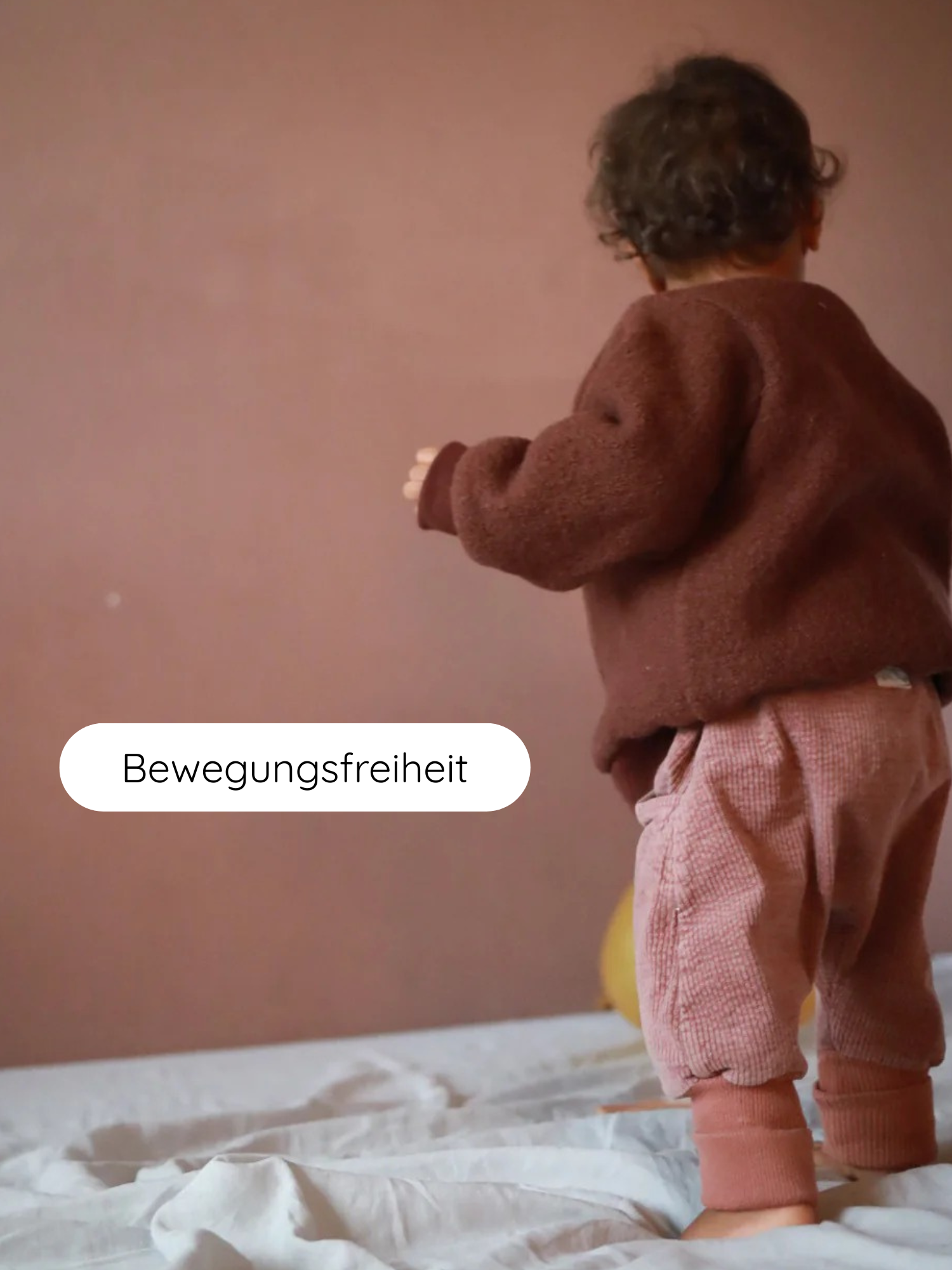 Kleines Kind auf weißem Bettlaken trägt Knickerbocker Breitcord Stretch - Rosenholz. Die lockeren, rose-pinkfarbenen Hosen bieten Bewegungsfreiheit. Im Hintergrund ein gelber Ballon, Wand in gedämpftem Rosé-Beige.