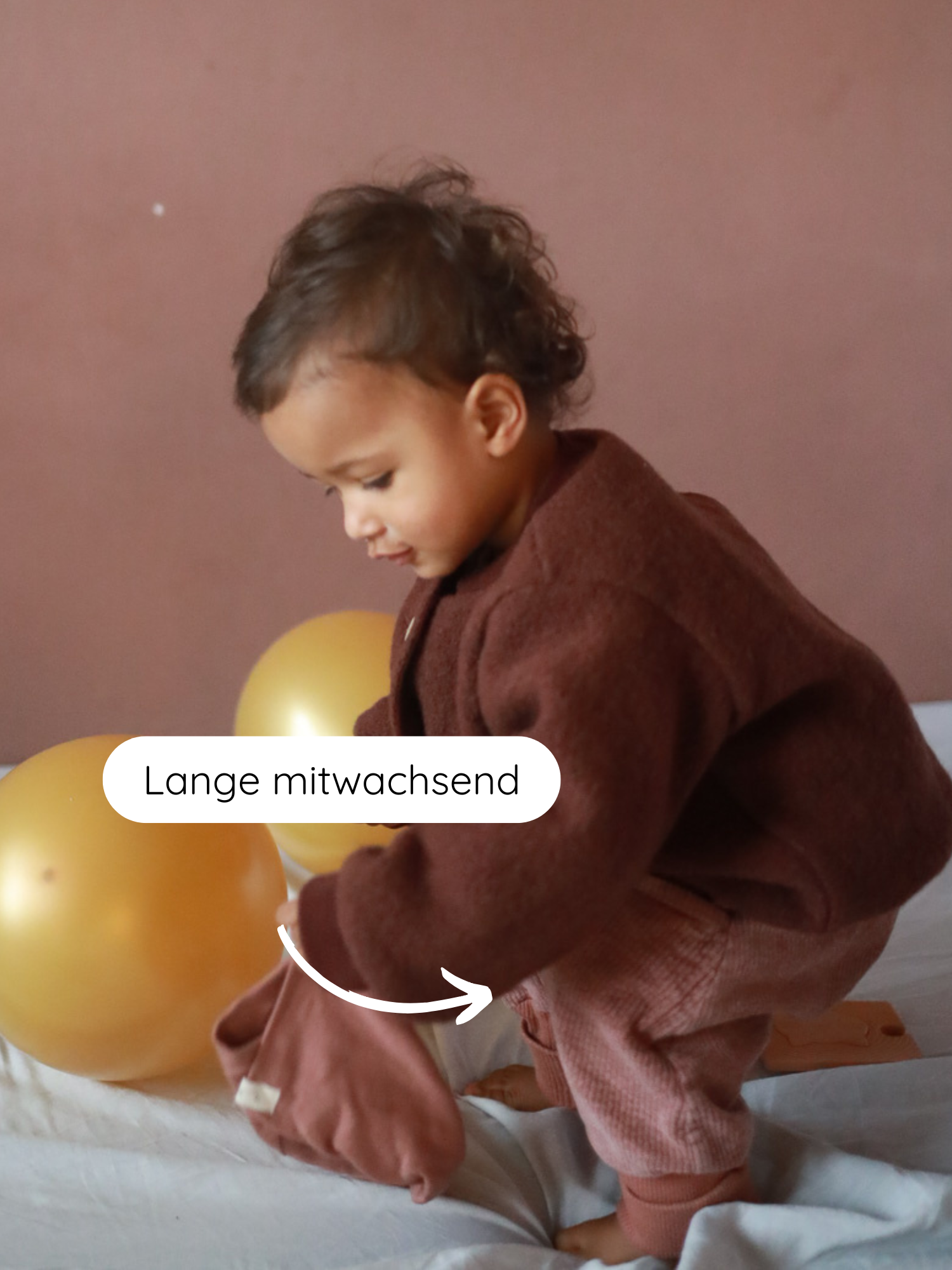 Kleinkind trägt Knickerbocker Breitcord Stretch - Rosenholz mit aufgerollten Bündchen, die mitwachsen. Neben ihm goldene Ballons, hält passende braune Kleidung; harmonisch in neutralem Raum.