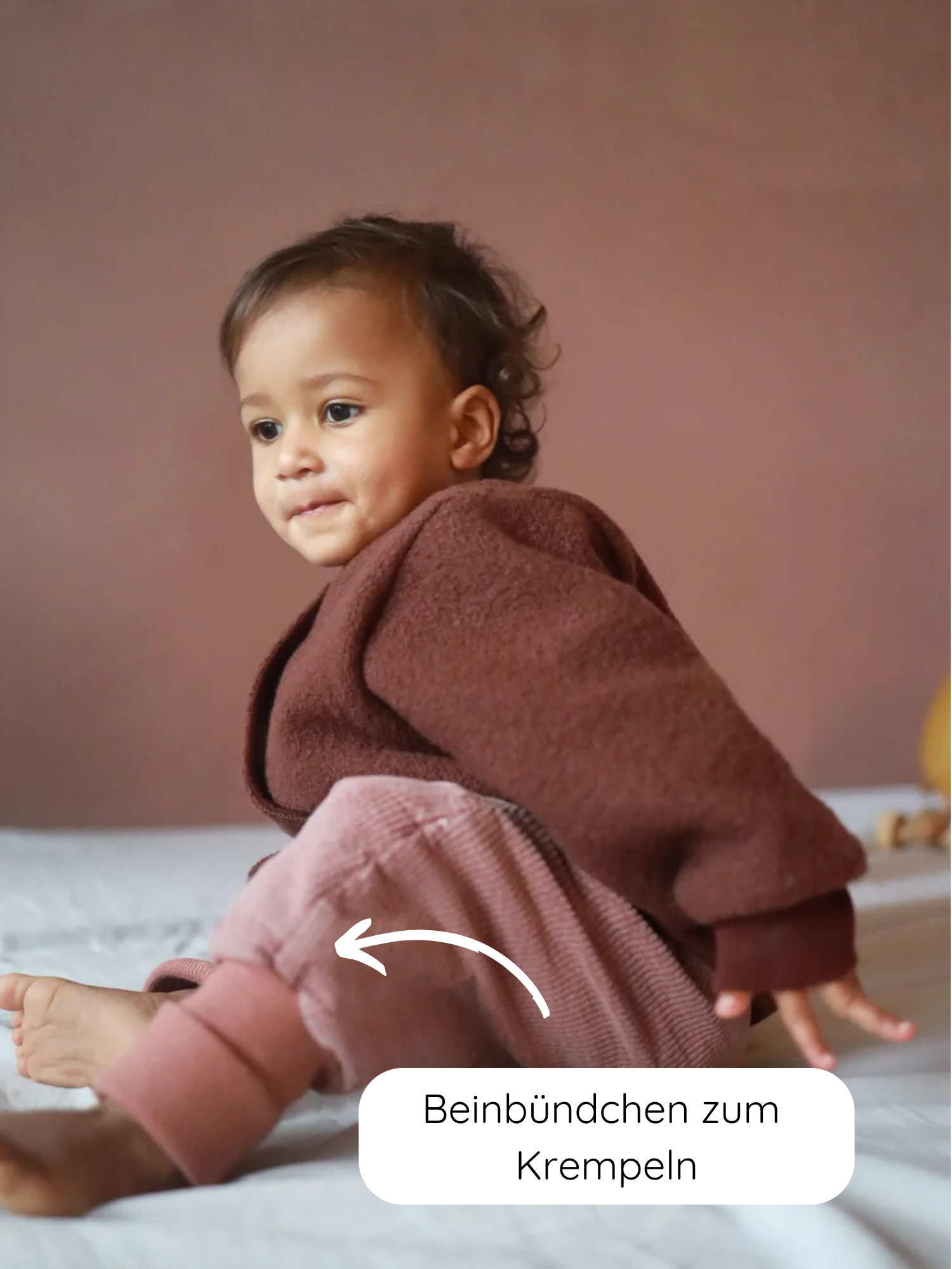 Kleinkind sitzt auf einer weichen Unterlage, trägt Knickerbocker Breitcord Stretch - Rosenholz mit aufrollbaren Beinbündchen, die Bewegungsfreiheit und Komfort für kühlere Tage bieten.