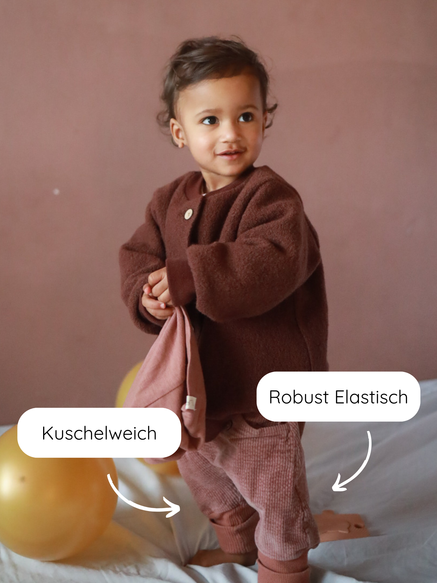 Kleinkind trägt Knickerbocker Breitcord Stretch - Rosenholz, kombiniert mit brauner Strickjacke. Die Hose bietet Bewegungsfreiheit und Komfort, perfekt für aktive Kinder. Neben dem Kind liegt ein goldener Luftballon.
