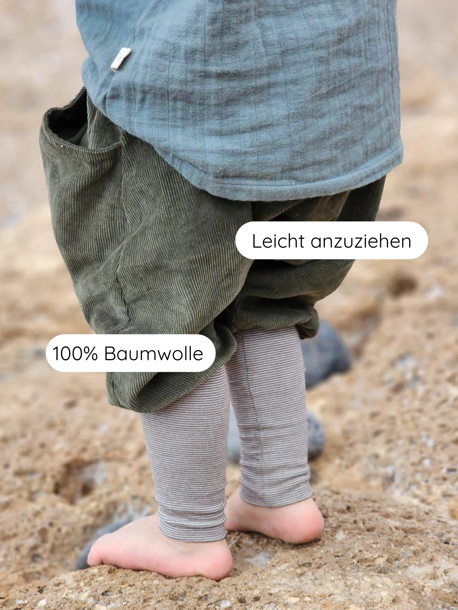 Kleinkind trägt Knickerbocker Feincord Ringel - Avocado, mit übergroßen olivgrünen Cordhosen über gestreiften Leggings, auf sandigem Untergrund. Perfekt für aktives Spielen, einfach anzuziehen, 100% Baumwolle.