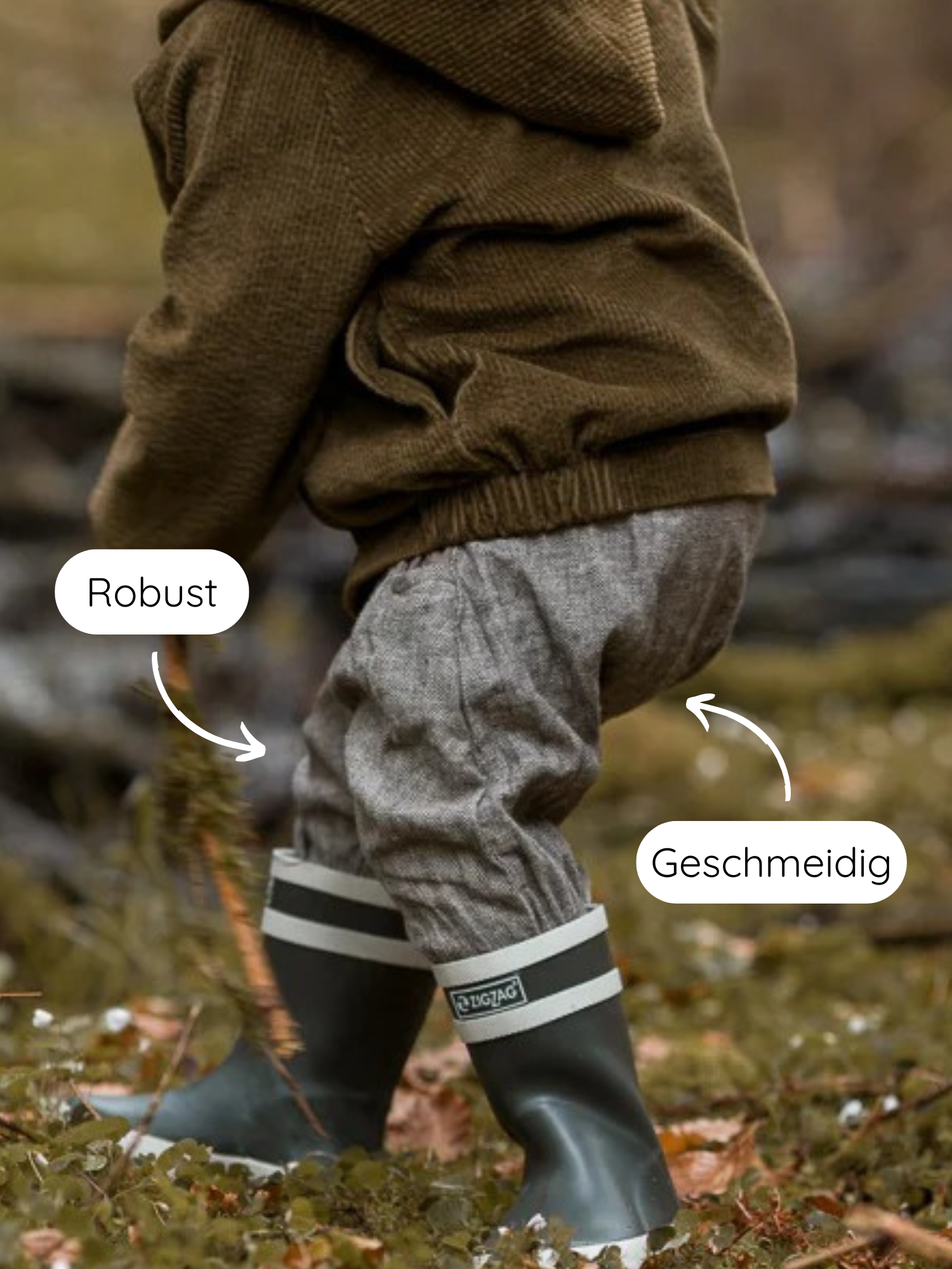 Knickerbocker Fischgrat: Ein Kind spielt draußen, trägt lockere, elastische graue Hosen aus Baumwoll-Leinen-Mix, perfekt für Bewegungsfreiheit, kombiniert mit robusten, olivgrünen Gummistiefeln und einem braunen Strickoberteil.