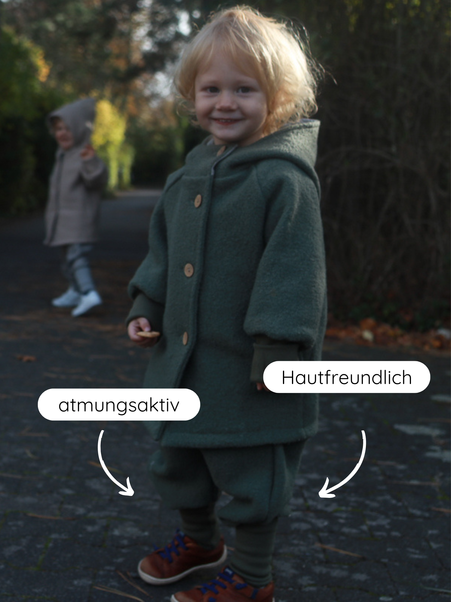 Kleinkind im Wollwalk Knickerbocker - Pistazie, mit passender Kapuzenjacke auf einem Weg, hält einen Keks. Textblase zeigt auf atmungsaktiven und hautfreundlichen Stoff, betont Materialqualität.