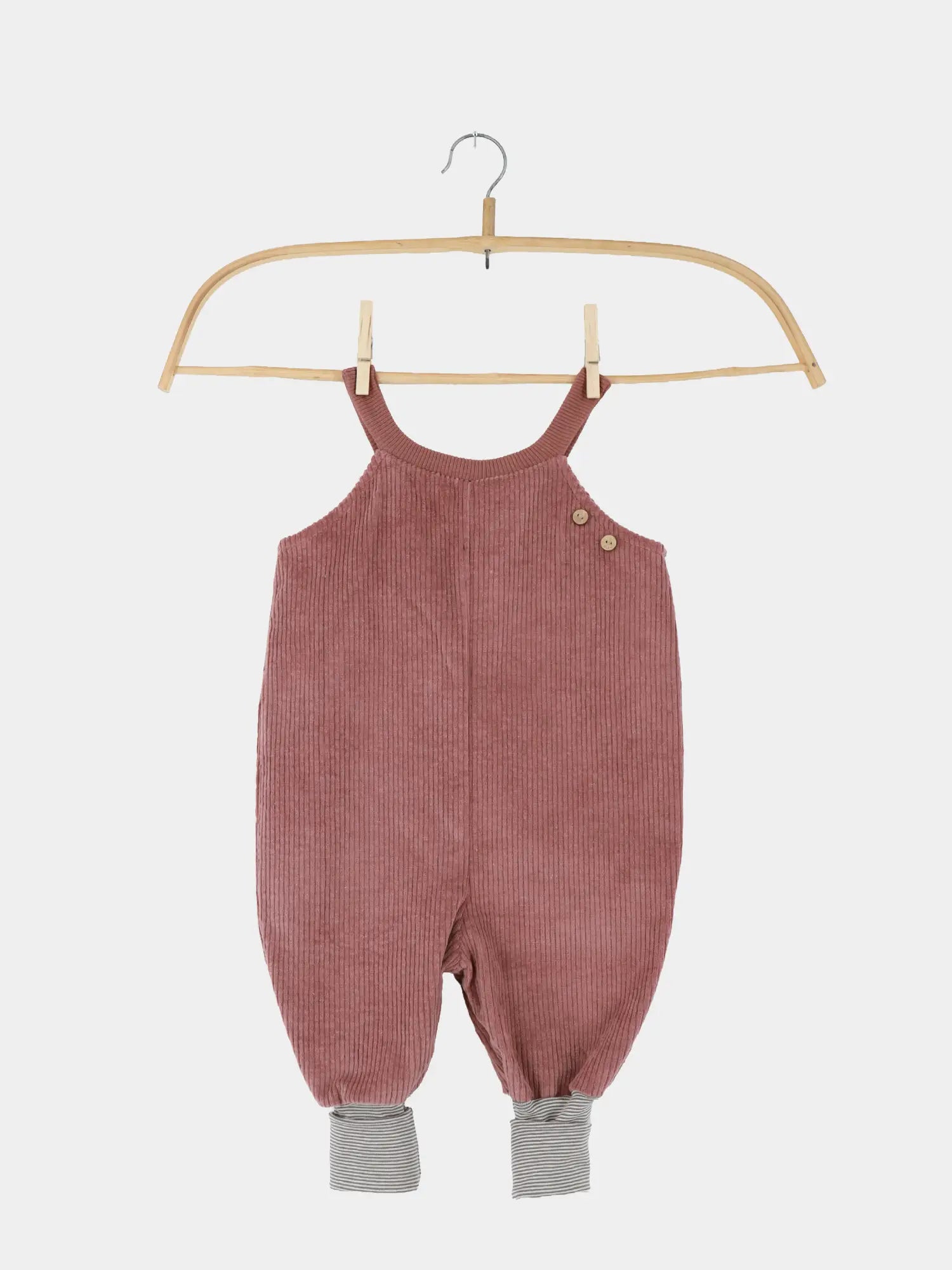 Schlupflatzhose Breitcord - Rosenholz: Ein Baby-Toddler-Romper aus staubrosa Kord, mit Holzknöpfen an Trägern und grau-weiß gestreiften Bündchen an den Hosenbeinen, auf einem Holzkleiderbügel hängend.