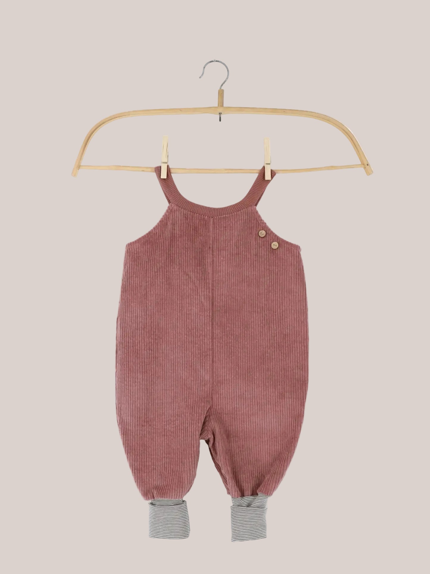 Schlupflatzhose Breitcord - Rosenholz: Ein Baby-Toddler-Romper aus staubrosa Kord, mit Holzknöpfen an Trägern und grau-weiß gestreiften Bündchen an den Hosenbeinen, auf einem Holzkleiderbügel hängend.