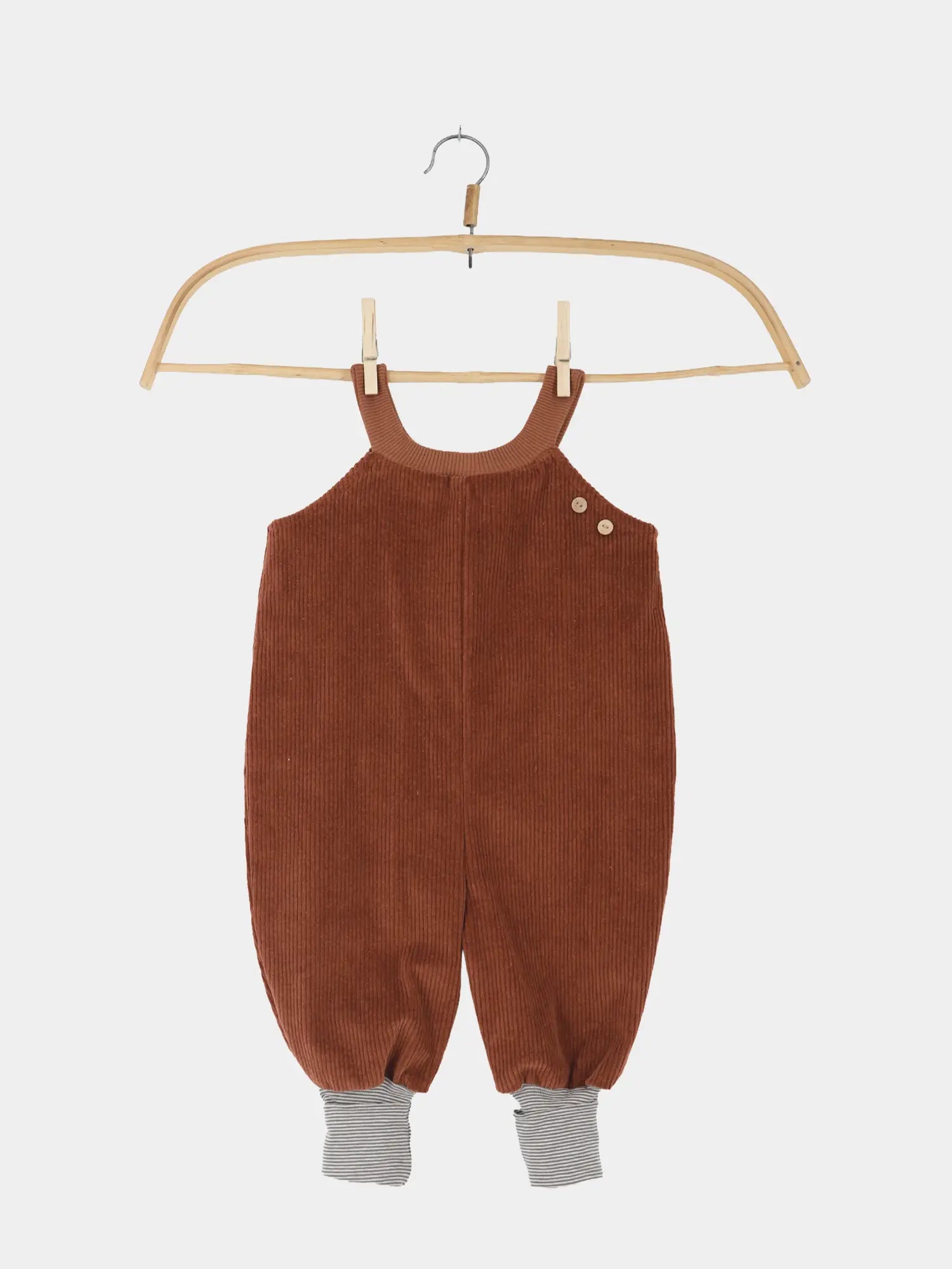 Schlupflatzhose Breitcord - Rost: Eine kleine braune Cord-Latzhose für Babys mit zwei Schulterträgern, Holzknöpfen, elastischen Beinabschlüssen und gestreiften Bündchen, hängend an einem Holzkleiderbügel.