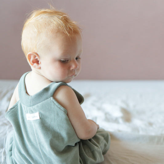 Kind sitzt auf Bett. Trägt Latzhose für Babys aus Baumwolle. Bequeme Kinderkleidung aus Berlin und Erstausstattung für Babys.