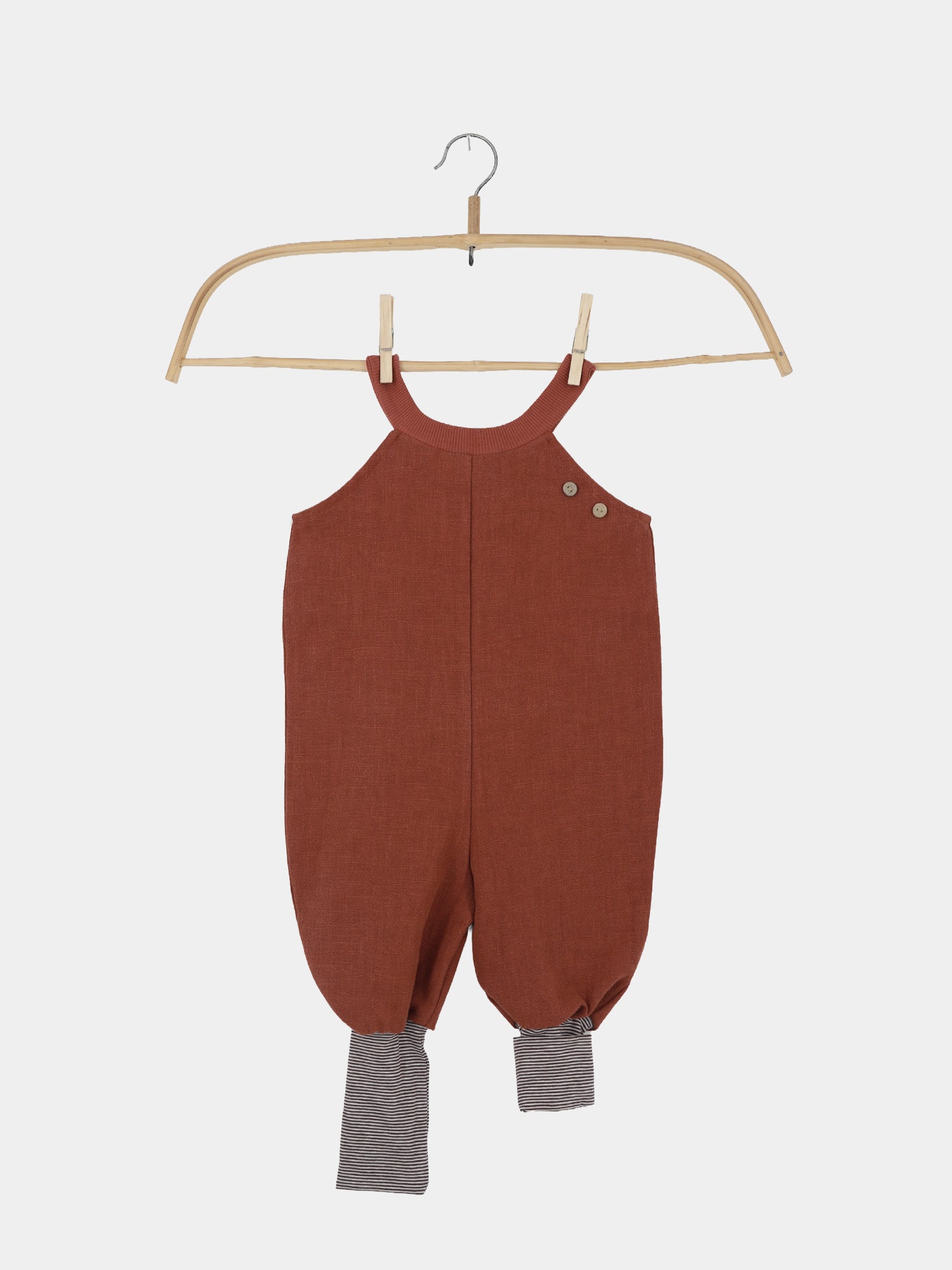 Schlupflatzhose Leinen - Ziegel: Rustbrauner, ärmelloser Babystrampler aus Leinen, mit zwei Knöpfen an der Schulter und gestreiften Bündchen an den Knöcheln, auf einem Holzkleiderbügel hängend.